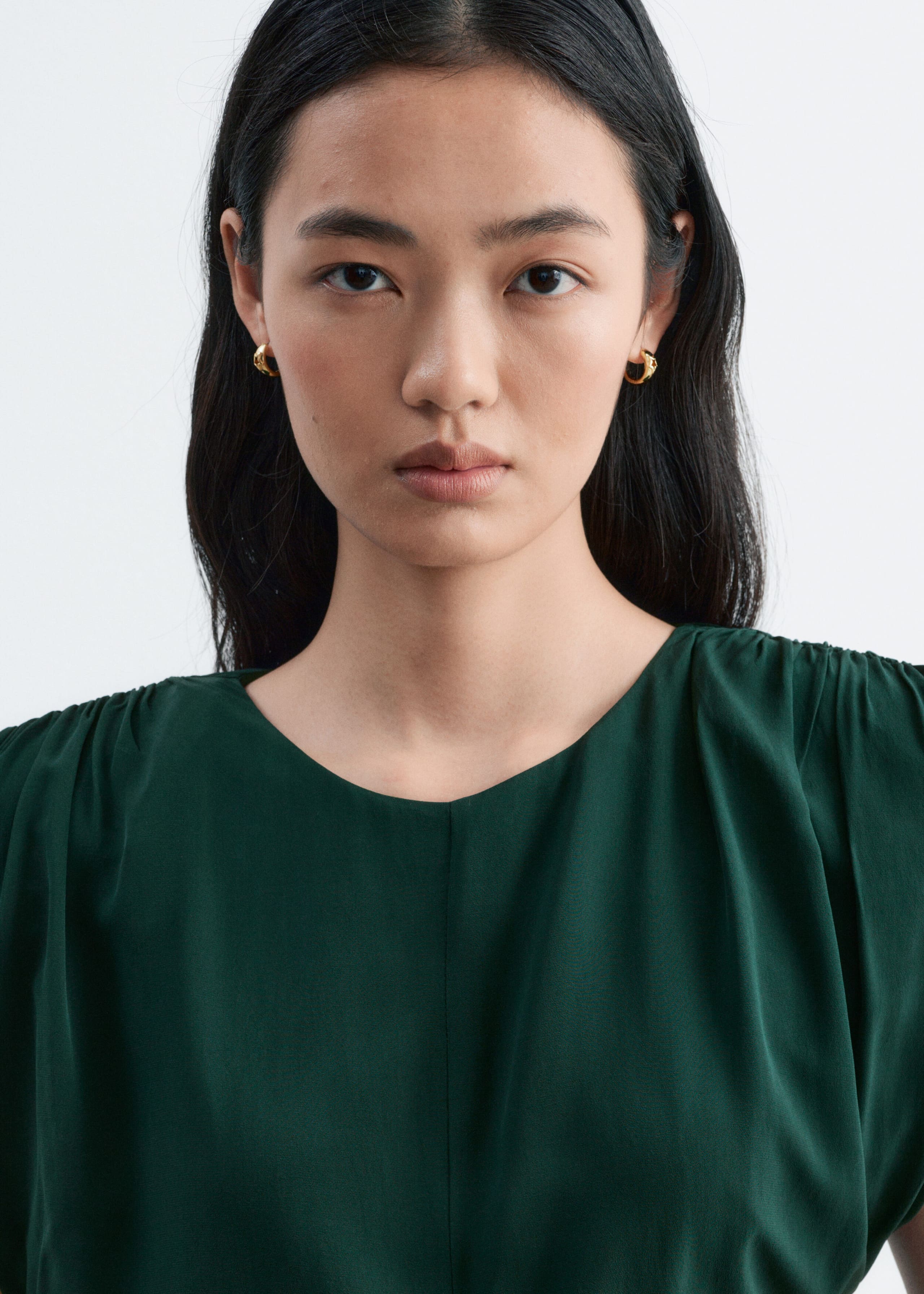 The model is wearing a dark green sleeveless top with a round neckline, a central vertical seam, and delicate ruching at the shoulders.