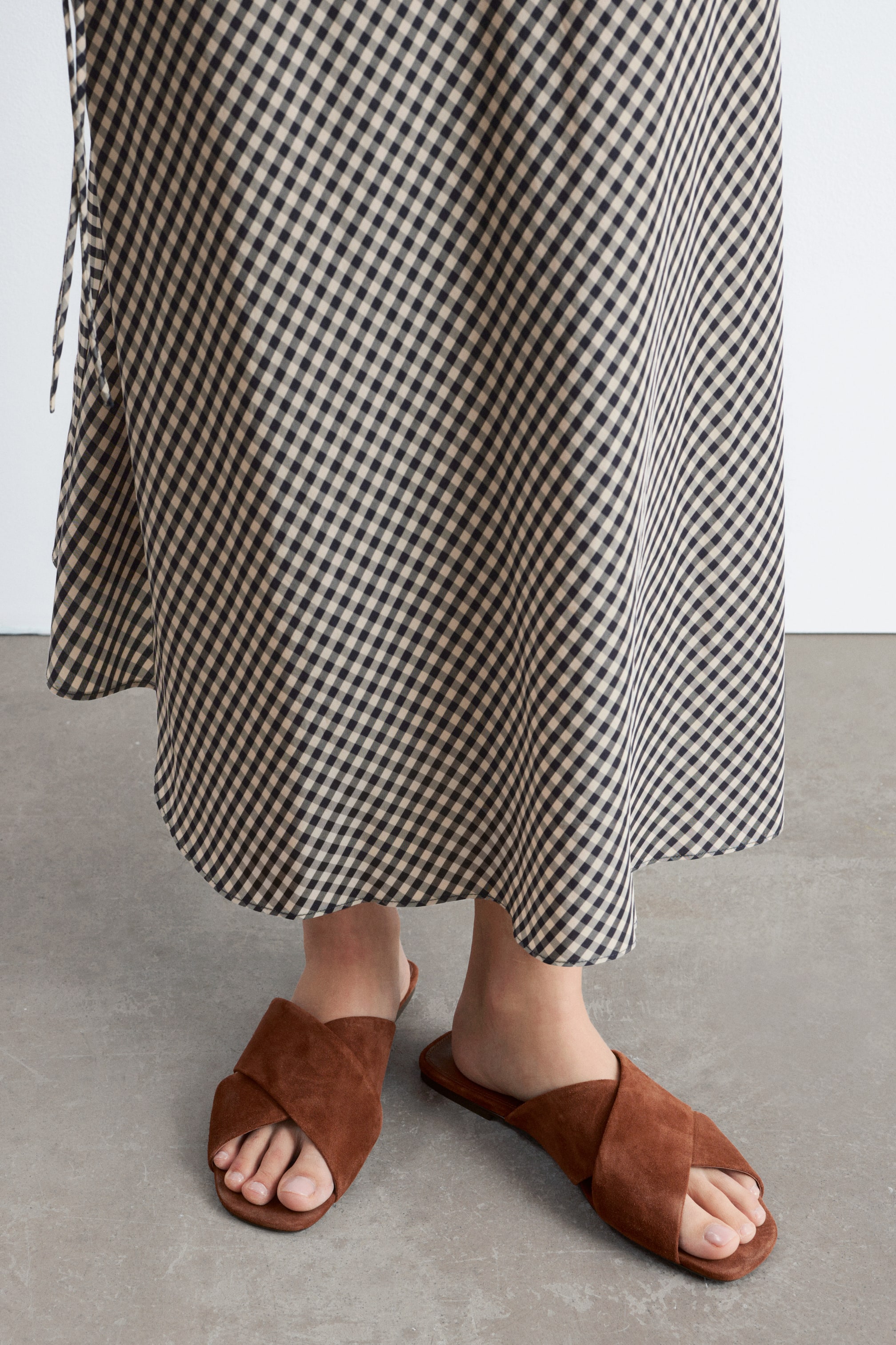 Ver imagem maior: Uma saia midi em xadrez vichy preto e bege flui em torno das pernas, apresentando um design transpassado com um detalhe de laço visível no lado esquerdo e uma bainha suavemente curvada.