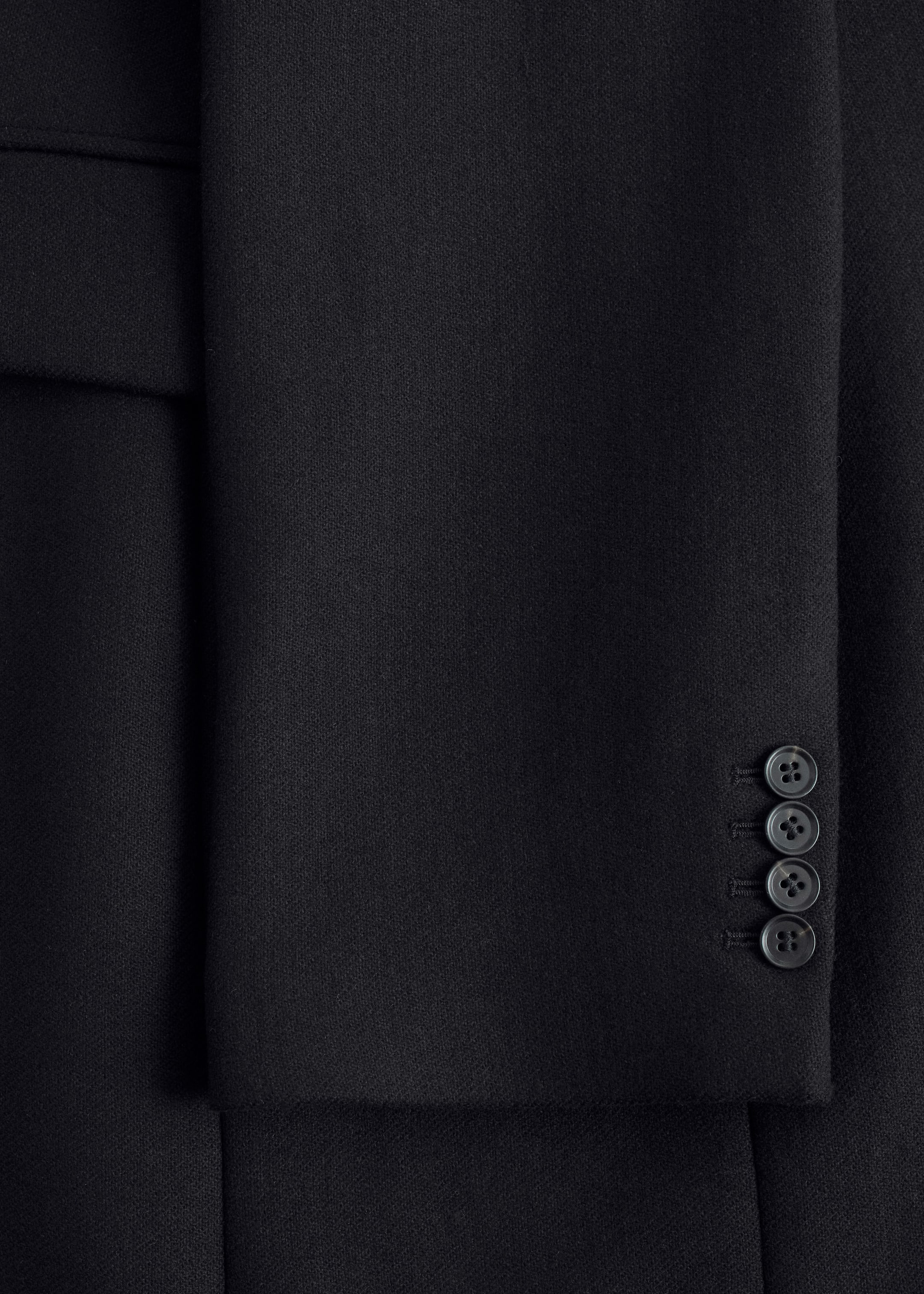 View larger image: A black wool-blend coat is featuring a subtle texture, visible in a close-up shot. A vertical fold is defining the left side of the garment, and the right sleeve cuff is displaying four dark, round buttons stacked vertically.