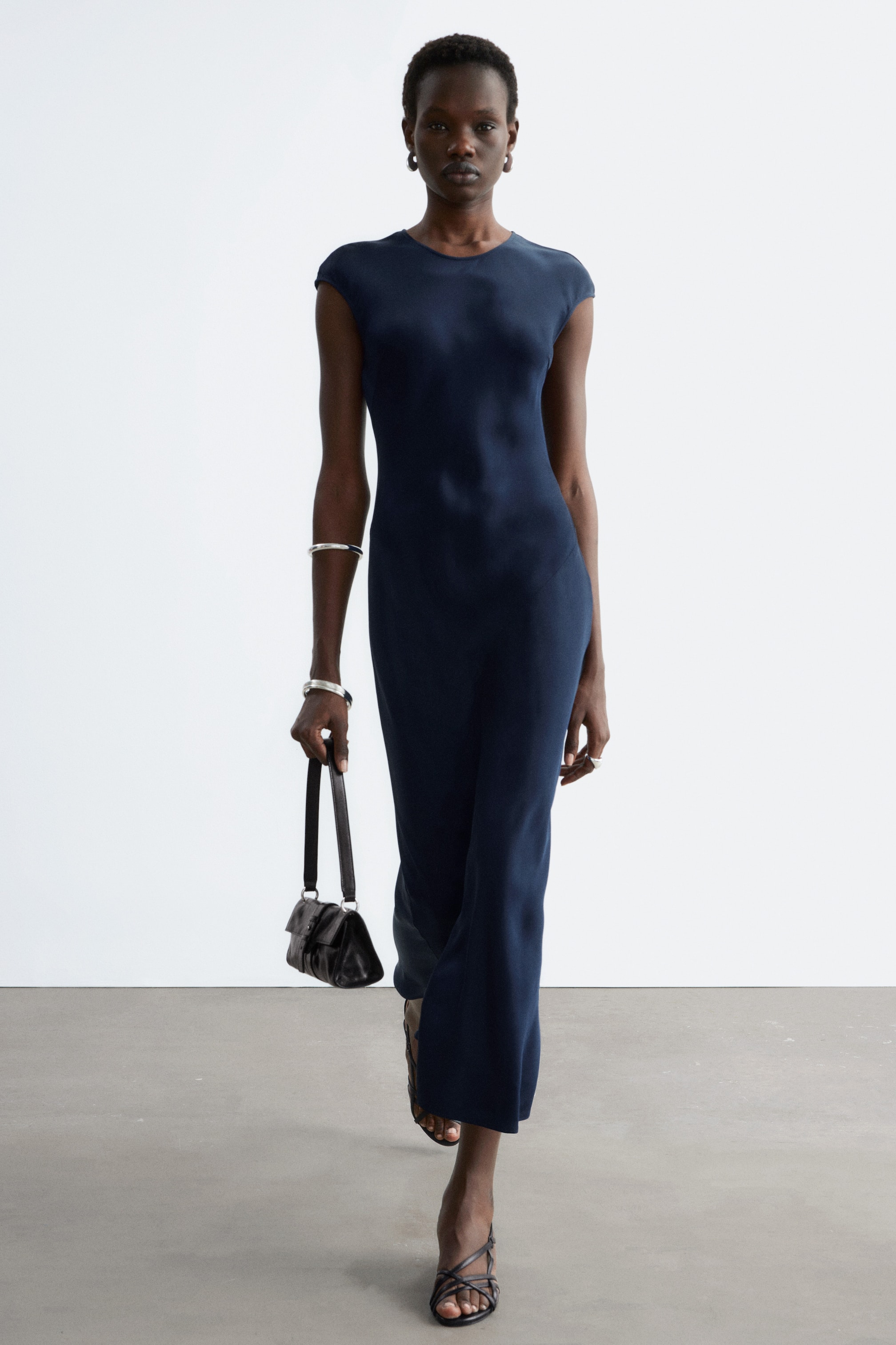 הצגת תמונה גדולה יותר: שמלה בצבע כחול כהה, עשויה מבד חלק ומבריק, מציגה צווארון עגול ושרוולים קצרים. היא מציגה צללית שנצמדת לגוף ומגיעה לאורך מידי עם שסע בצד.