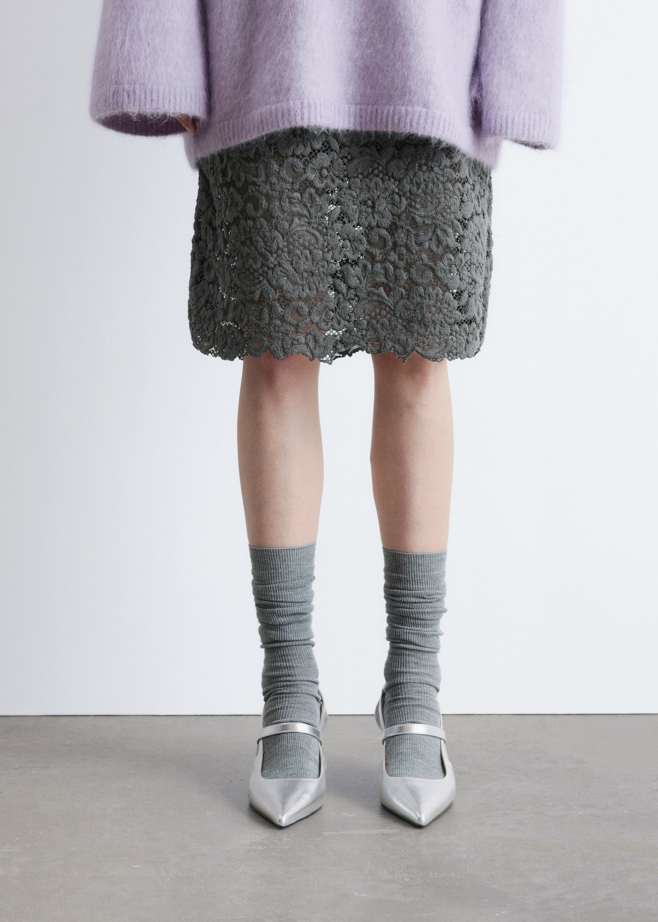 Agrandir l'image: Une jupe en dentelle grise est présentée, avec un motif floral compliqué et un ourlet festonné. Elle tombe juste au-dessus des genoux et est superposée sous un pull violet clair.