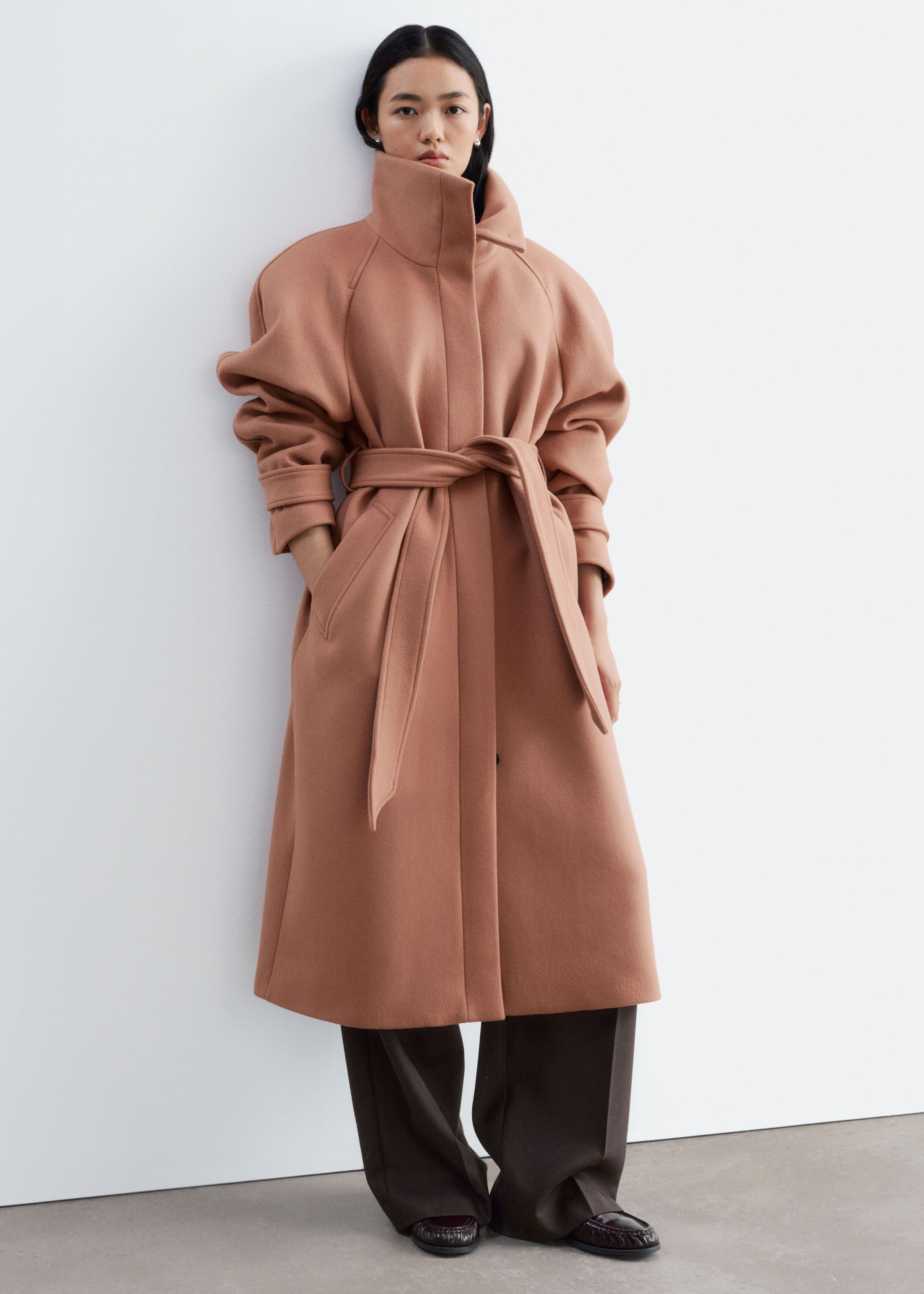 A rose-brown wool-blend coat featuring a high funnel neck, rounded shoulders, and a matching tie belt is worn by the model. The model is standing with one hand in the side pocket, styling the coat with dark brown wide-leg trousers.