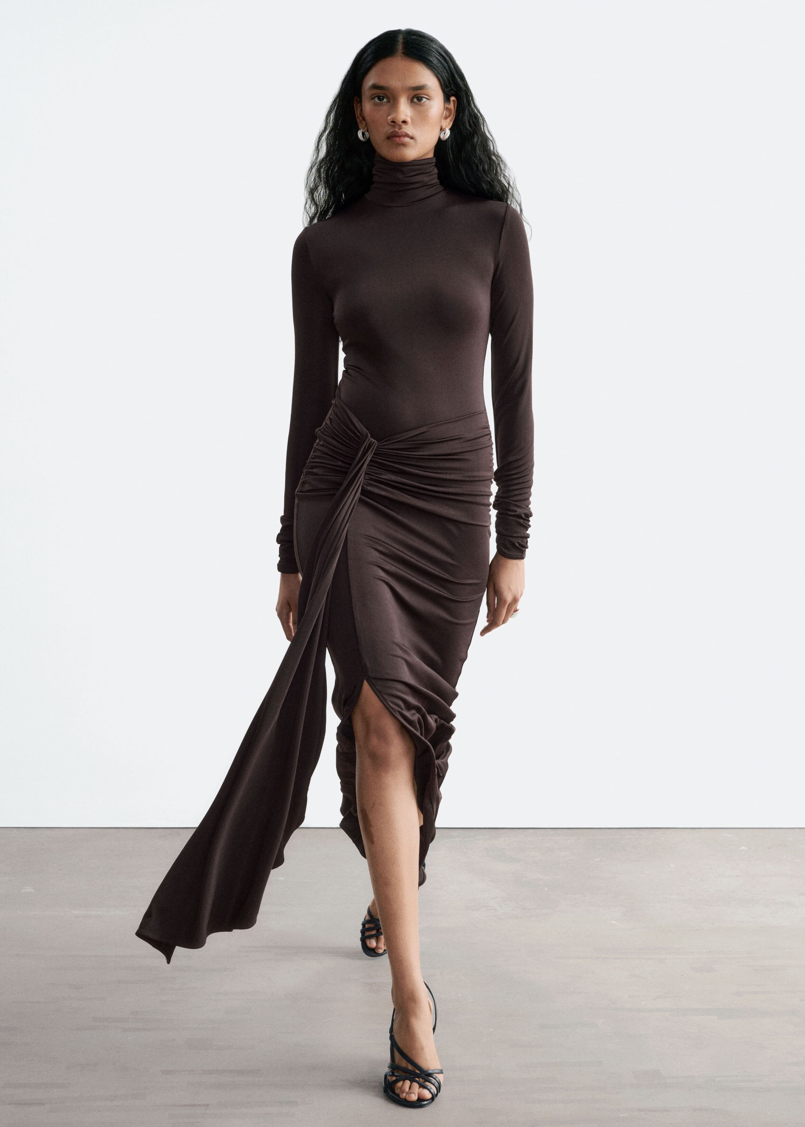 The model is wearing a dark brown jersey dress featuring a slim fit, long sleeves and a mock neck. The dress is designed with a draped tie detail at the waist that falls into a long fabric panel, revealing a front slit as the model steps forward.