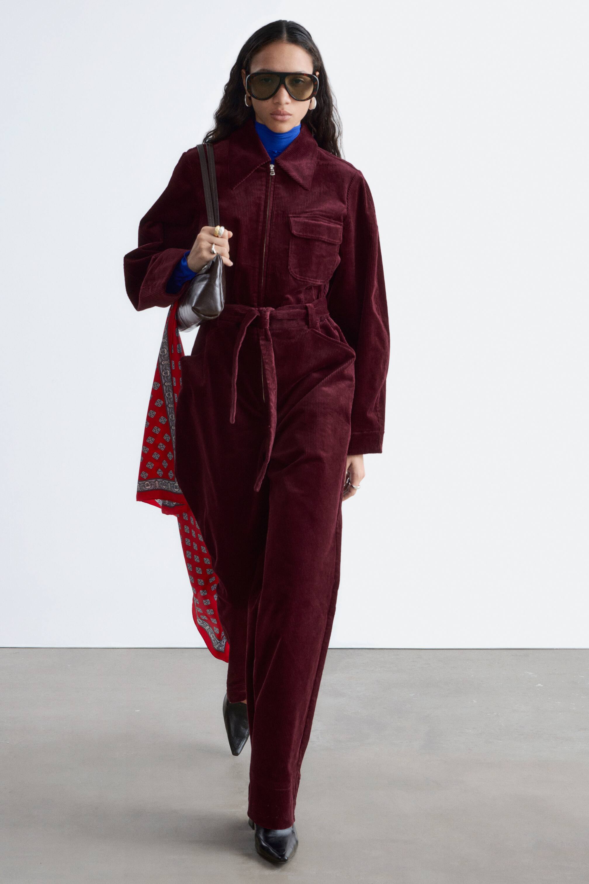 Visualizza immagine più grande: Una tuta in velluto a coste bordeaux presenta un colletto a punta, una zip frontale a tutta lunghezza, due tasche sul petto con patta e una cintura in vita con nodo che stringe il punto vita. È caratterizzata da pantaloni a gamba larga ed è stratificata su un dolcevita blu, con una sciarpa a fantasia rossa infilata di lato.