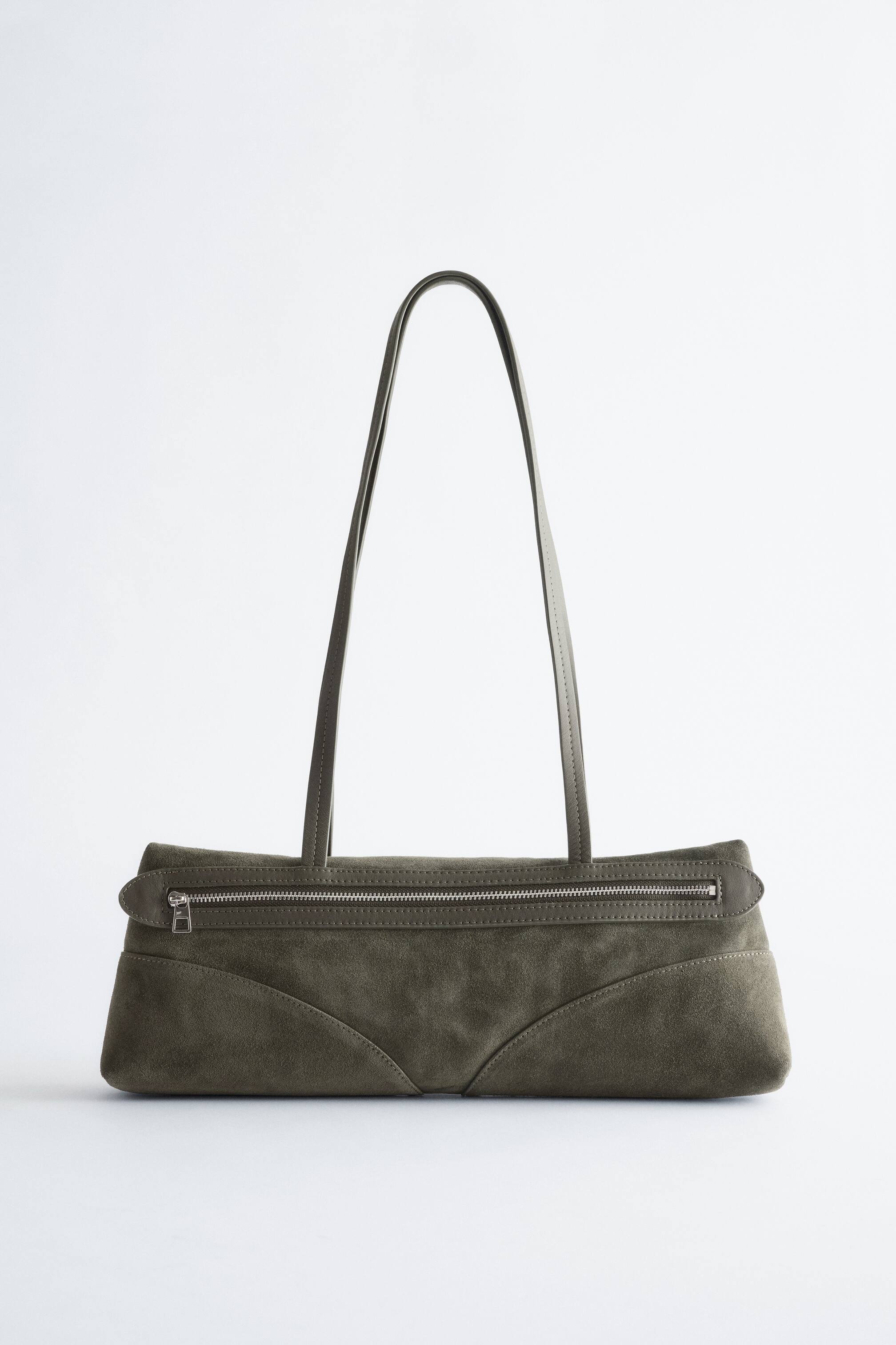 View larger image: A low-profile, rectangular shoulder bag is presented FRONT, featuring a muted green suede body with curved seam details on the lower front. It is displaying a silver-toned zip closure along the top and long, thin green leather shoulder straps.