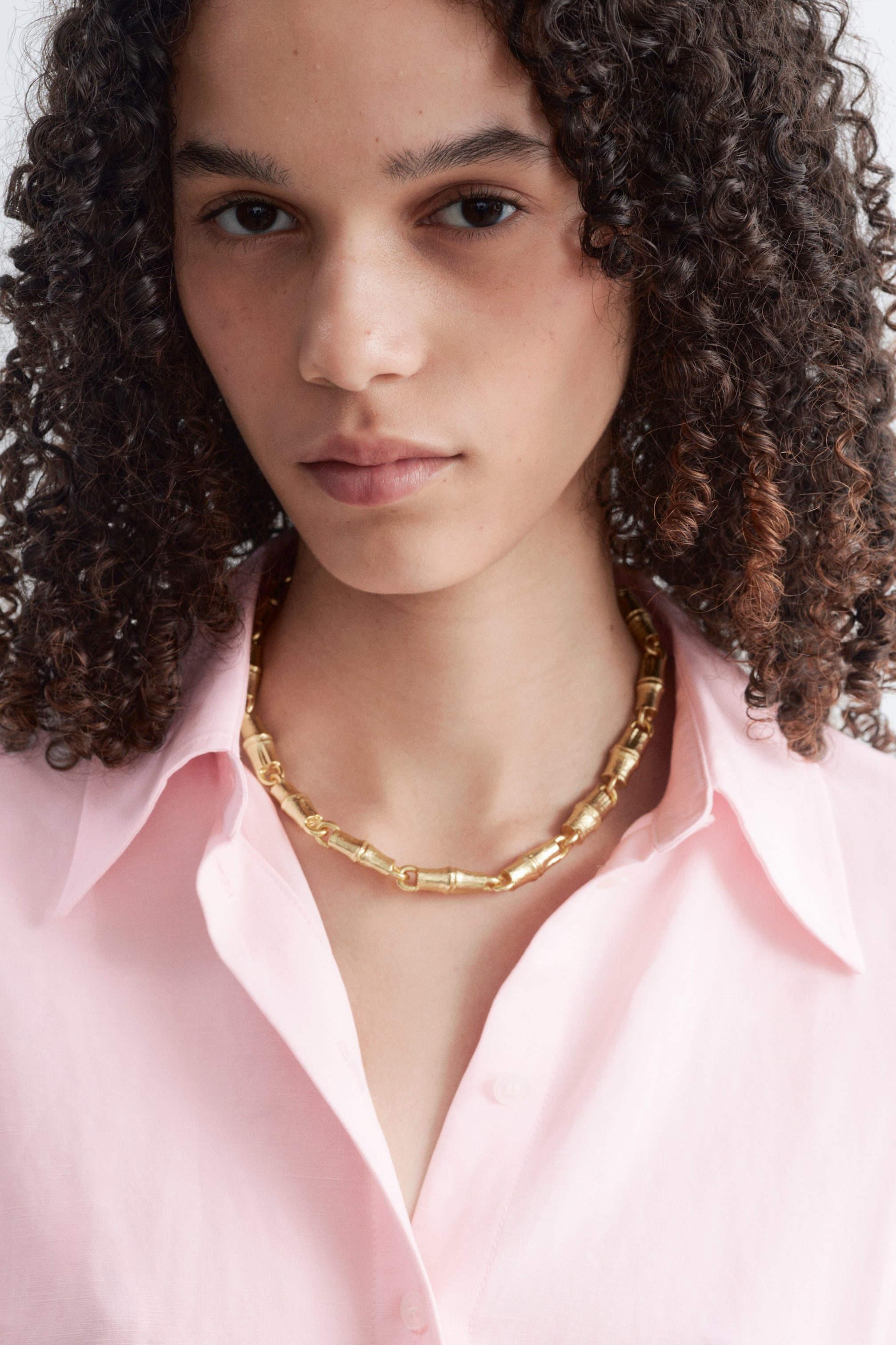 Visualizza l'immagine ingrandita: Una collana a catena dorata con texture, caratterizzata da maglie cilindriche segmentate simili al bambù, indossata dalla modella. La collana poggia sulla clavicola ed è abbinata a una camicia rosa con colletto aperto.