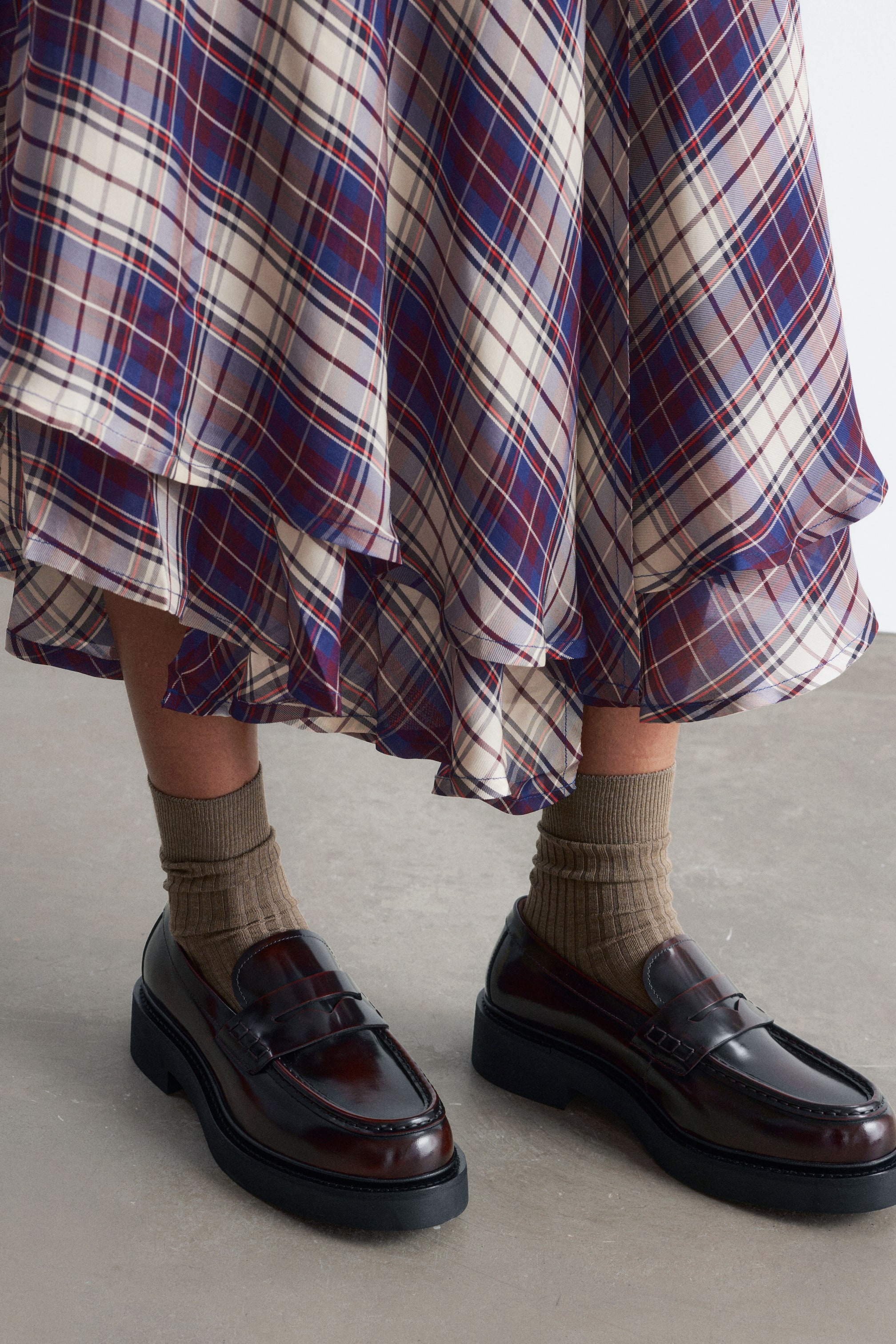 Ver imagen más grande: Unas bailarinas tipo penny en cuero burdeos oscuro de alto brillo, con detalles de pespunte y una suela gruesa de goma negra, están siendo lucidas por la modelo. Los zapatos se combinan con calcetines beige de canalé y una falda de cuadros en capas.