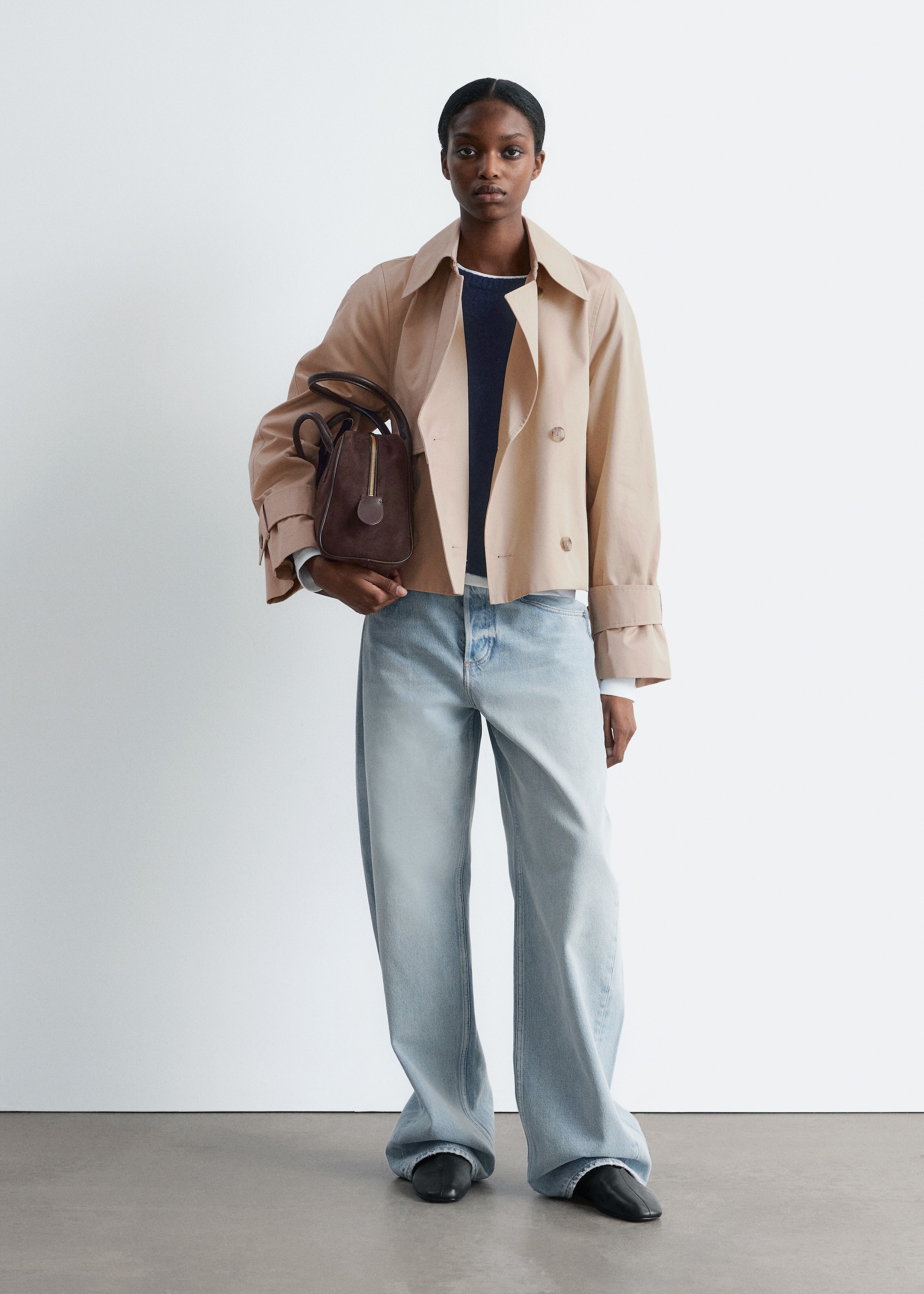 Größeres Bild ansehen: Ein Model präsentiert einen kurzen, beigen, zweireihigen Trenchcoat mit breiten Revers und sichtbaren Manschettenriemen, der offen über einem marineblauen Oberteil und einem weißen Langarmhemd getragen wird. Sie kombiniert dies mit hell gewaschenen, weit geschnittenen Jeans, die an den Knöcheln aufliegen, schwarzen geschlossenen Schuhen und hält eine dunkelbraune Handtasche mit einem auffälligen vertikalen Frontreißverschluss.