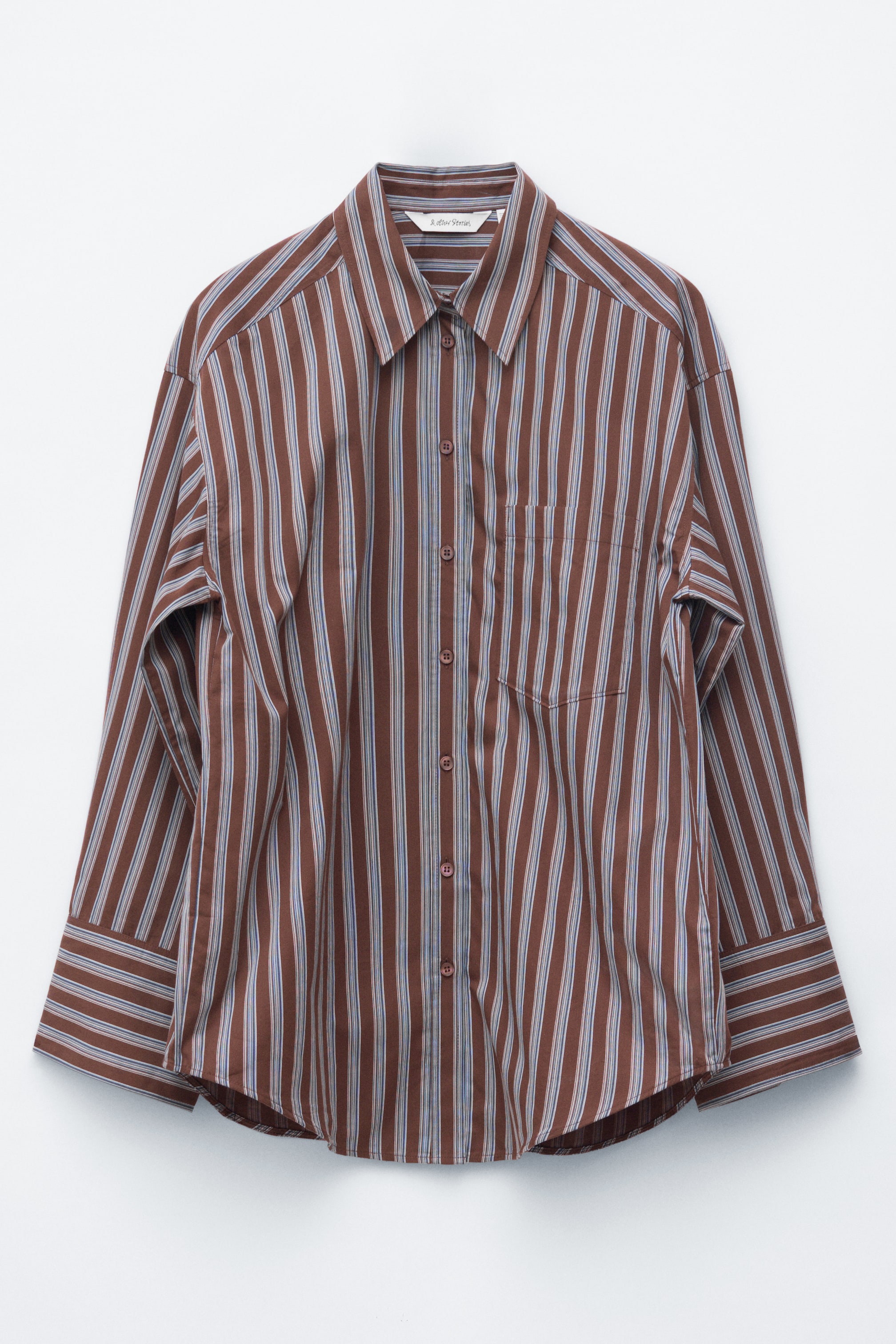 Visualizza immagine più grande: La camicia a maniche lunghe è presentata in vista FRONTALE, decorata con strisce verticali marroni, azzurro chiaro e sottili strisce bianche. È progettata con un colletto a punta, una paramontura a tutta lunghezza con bottoni marroni in tinta, una tasca applicata sul petto sinistro, polsini con bottoni e un orlo curvo.