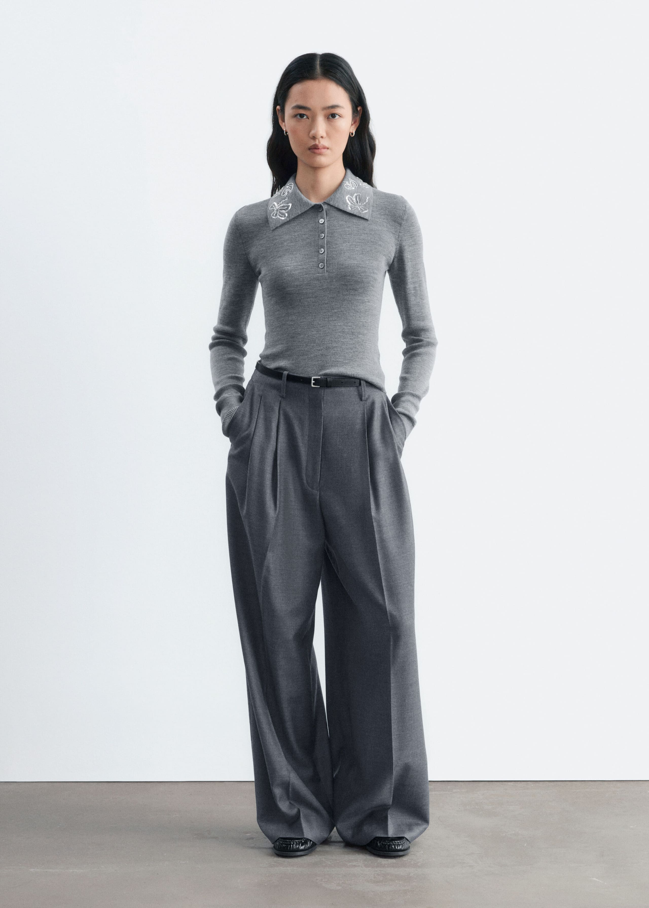 Ein grauer Woll-Polopullover mit Slim Fit, langen Ärmeln, einer geknöpften Frontleiste und weißen bestickten Designs am Kragen wird vom Model getragen. Der Pullover ist in eine weit geschnittene graue Hose gesteckt und mit einem dünnen schwarzen Gürtel gestylt.