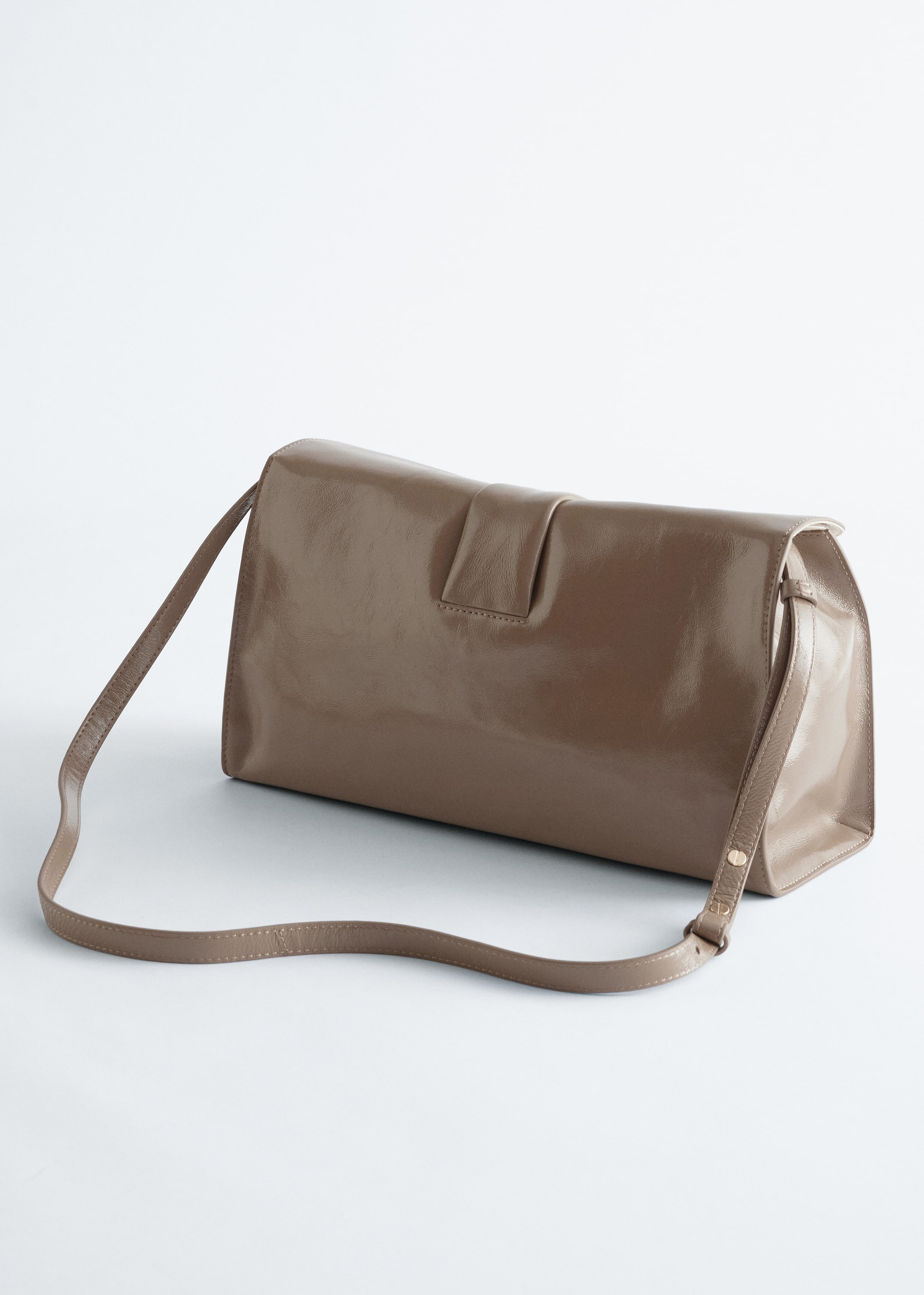 Größeres Bild ansehen: Eine glänzende, taupefarbene Leder-Clutch wird aus einem seitlichen und rückwärtigen Winkel angezeigt, mit einer oberen Klappe und einer zentralen Lasche. Ein dünner, verstellbarer Schulterriemen erstreckt sich von ihren Seiten.