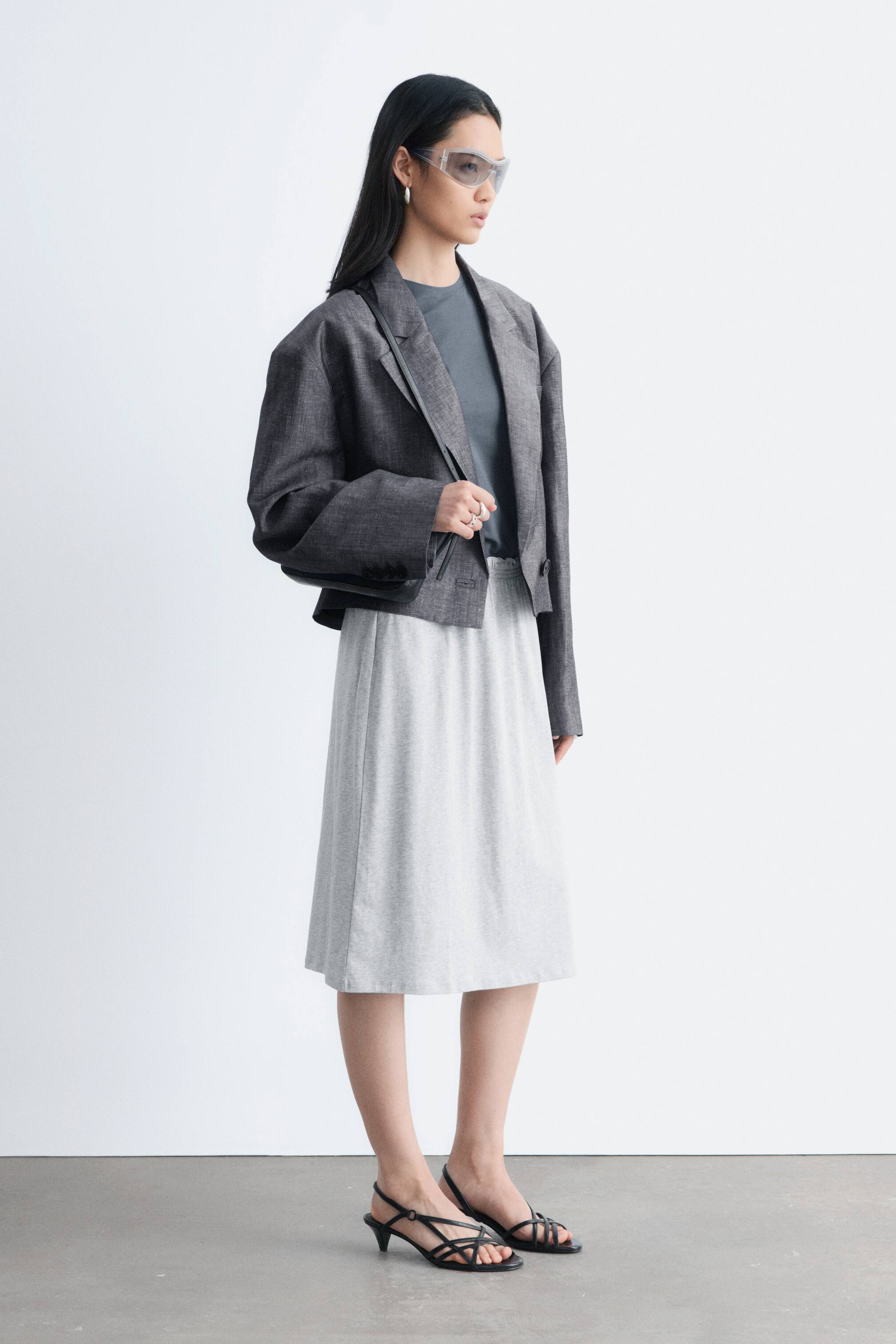 Visualizza immagine più grande: Una gonna grigia chiara al ginocchio, apparentemente realizzata in tessuto jersey, è indossata dalla modella. Presenta una cintura elastica arricciata ed è abbinata a una t-shirt grigio scuro e a un blazer corto.
