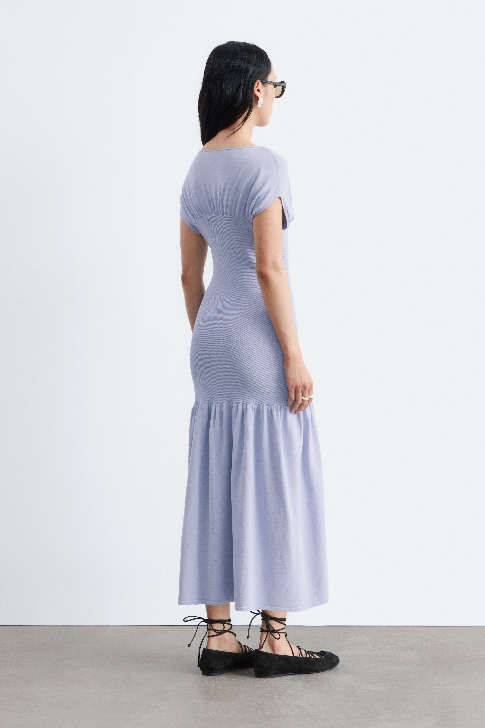 Ver imagen más grande: Se presenta un vestido color periwinkle desde un ángulo trasero-derecho. Presenta un diseño ajustado con textura acanalada, mangas cortas y fruncido en la parte superior de la espalda, que se transforma en un suave y fluido volante recogido desde la mitad de la pantorrilla hasta los tobillos.