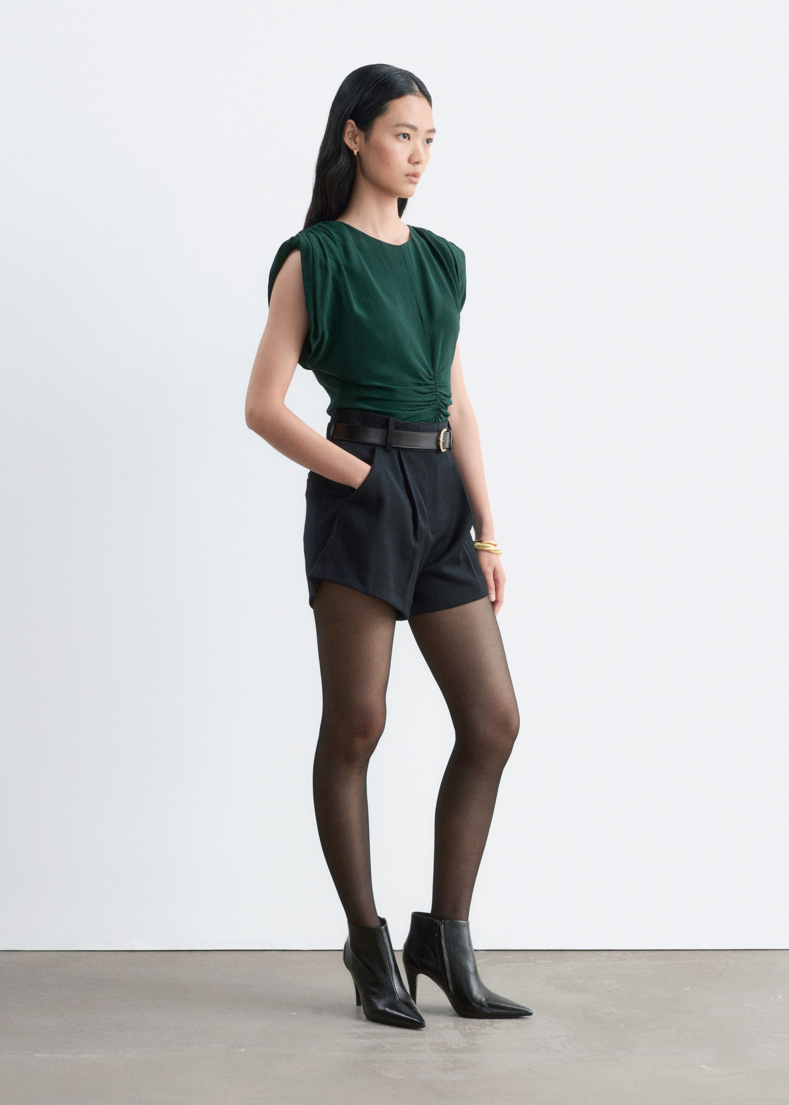 A dark green sleeveless top with ruching at the shoulders and center front is worn by the model. The top is tucked into high-waisted black shorts with a belt and styled with black tights and high-heeled ankle boots.