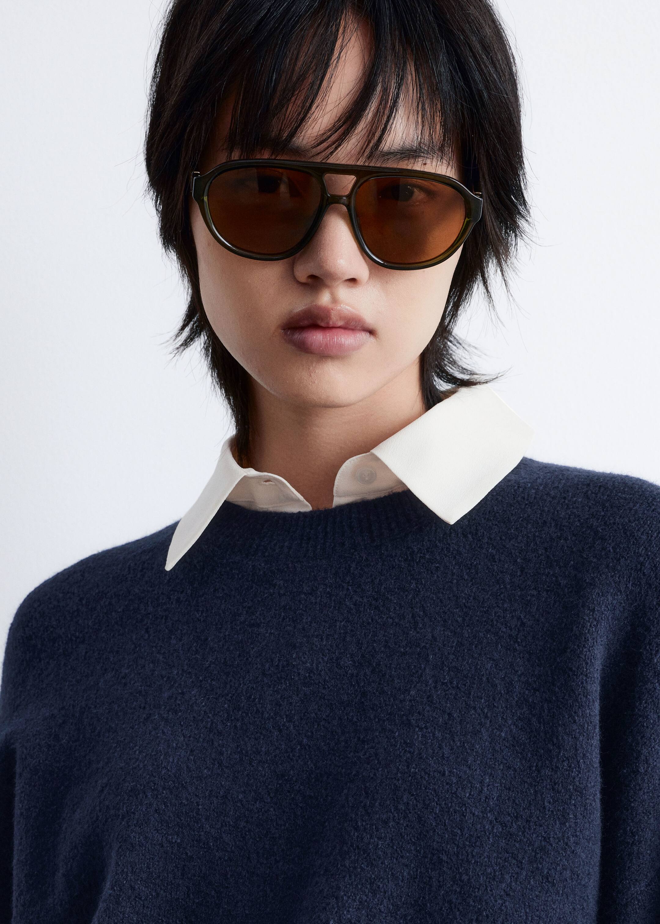Grösseres Bild ansehen: Dunkel olivgrüne Aviator-Sonnenbrillen mit braunen Gläsern werden getragen, leicht nach unten gerichtet. Ein flauschiger, marineblauer Pullover mit Rundhalsausschnitt ist sichtbar, über einem weißen Hemd mit Kragen geschichtet.