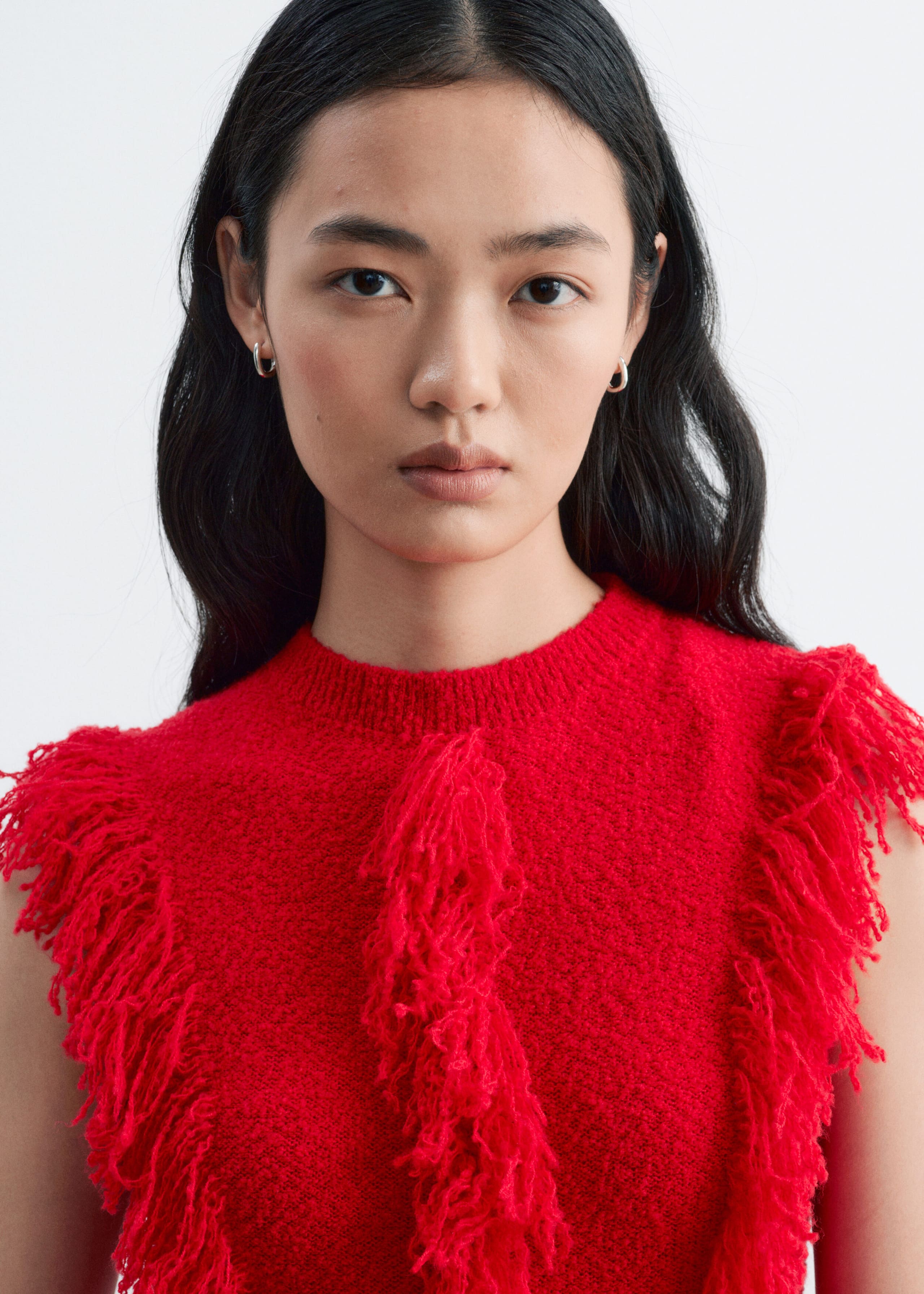 The model is wearing a red sleeveless knitted dress featuring a ribbed crew neckline and dense fringe detailing around the armholes and down the centre of the chest.