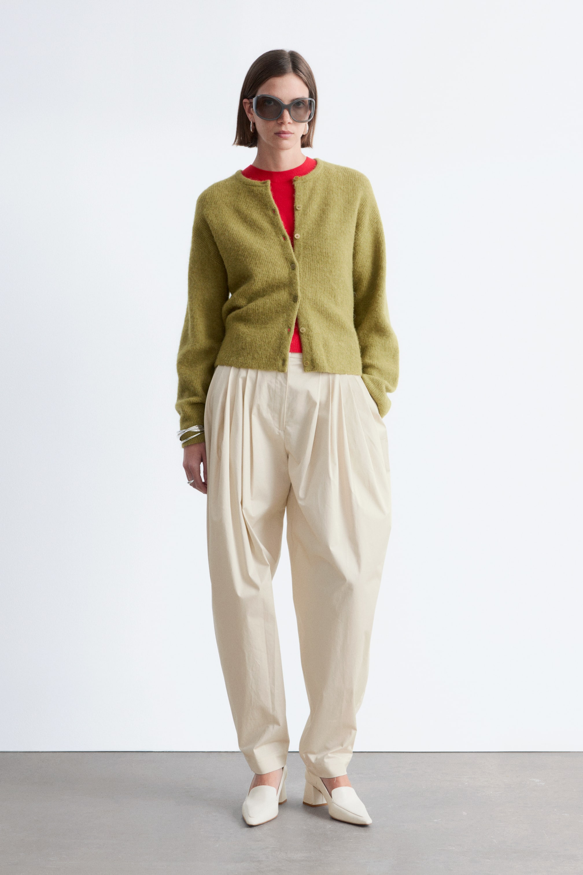 Grotere afbeelding bekijken: Een olijfgroene pluizige gebreide cardigan, met een ronde hals en lichtgekleurde knopen aan de voorkant, wordt gedragen over een rode top met ronde hals. Crèmekleurige geplooide broek met wijde pijpen wordt gecombineerd met puntige muiltjes met blokhakken in een bijpassende crèmekleur.