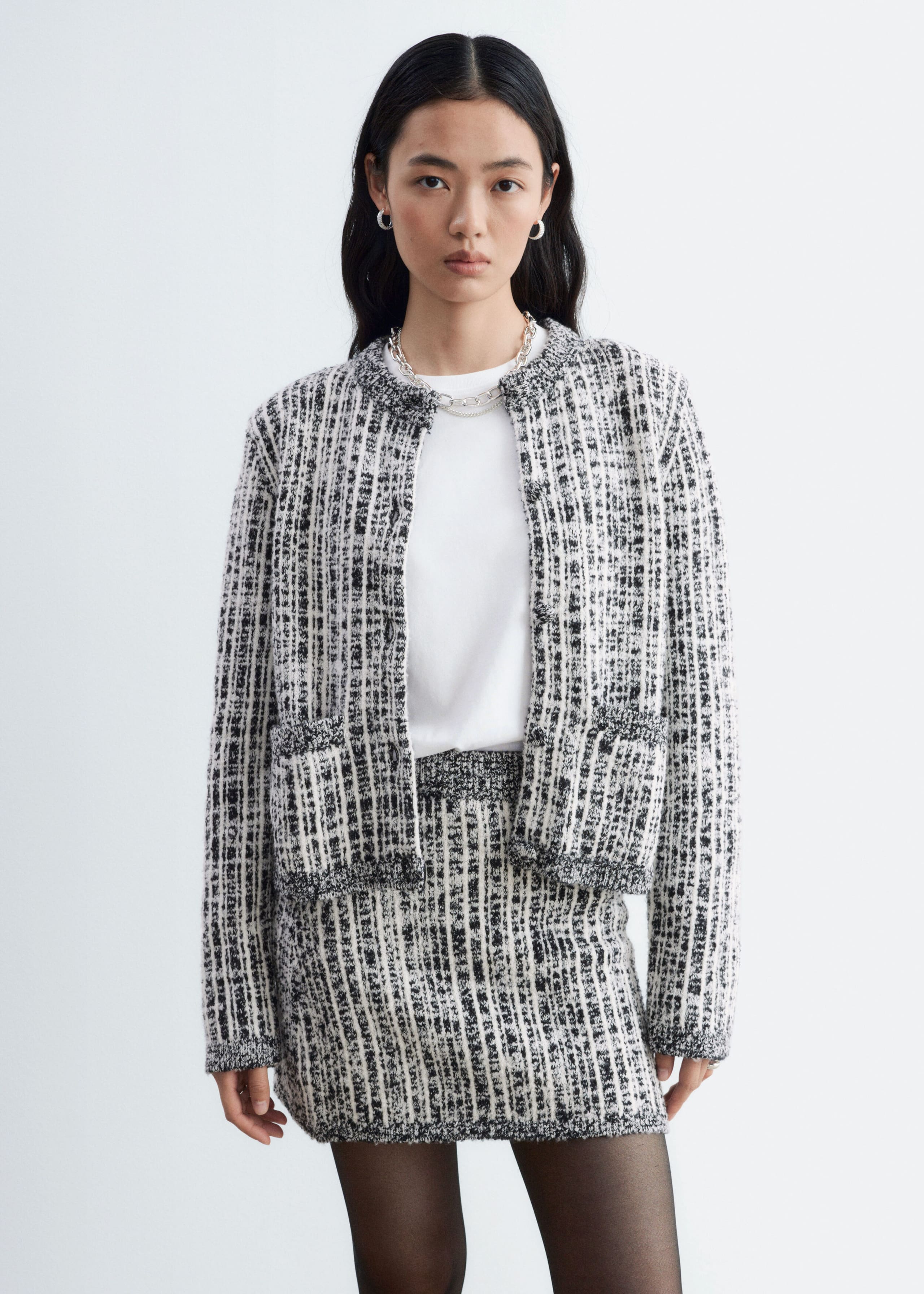 The model is wearing a textured black-and-white knit jacket with a matching mini skirt. The open-front, collarless jacket features front patch pockets and is layered over a white crewneck T-shirt, while the matching skirt is styled with sheer black tights.
