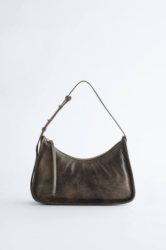Un sac à bandoulière en forme de croissant est présenté de FACE, fabriqué dans un matériau sombre, moucheté, à l'aspect vieilli. Il se ferme avec une fermeture éclair de couleur dorée le long de son sommet incurvé, sur laquelle pend une languette assortie, et est doté d'une fine bandoulière ajustable avec de petits éléments en métal doré. - 1