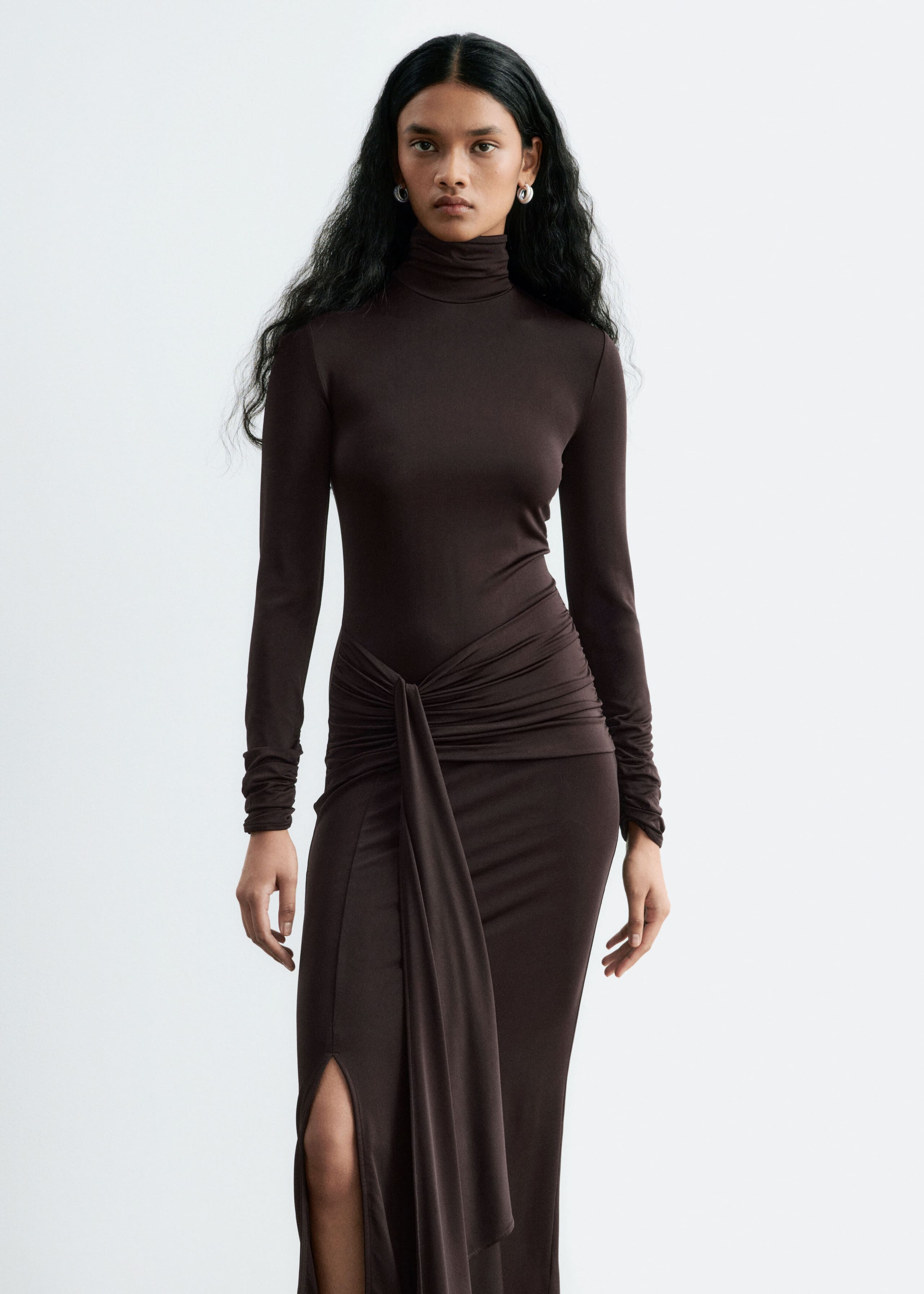 The model is wearing a dark brown jersey dress, featuring a double-layer mock neck, slim long sleeves, and a draped tie detail at the waist. The long-silhouette dress is styled with a gathered front panel that drapes down, alongside a high thigh split.