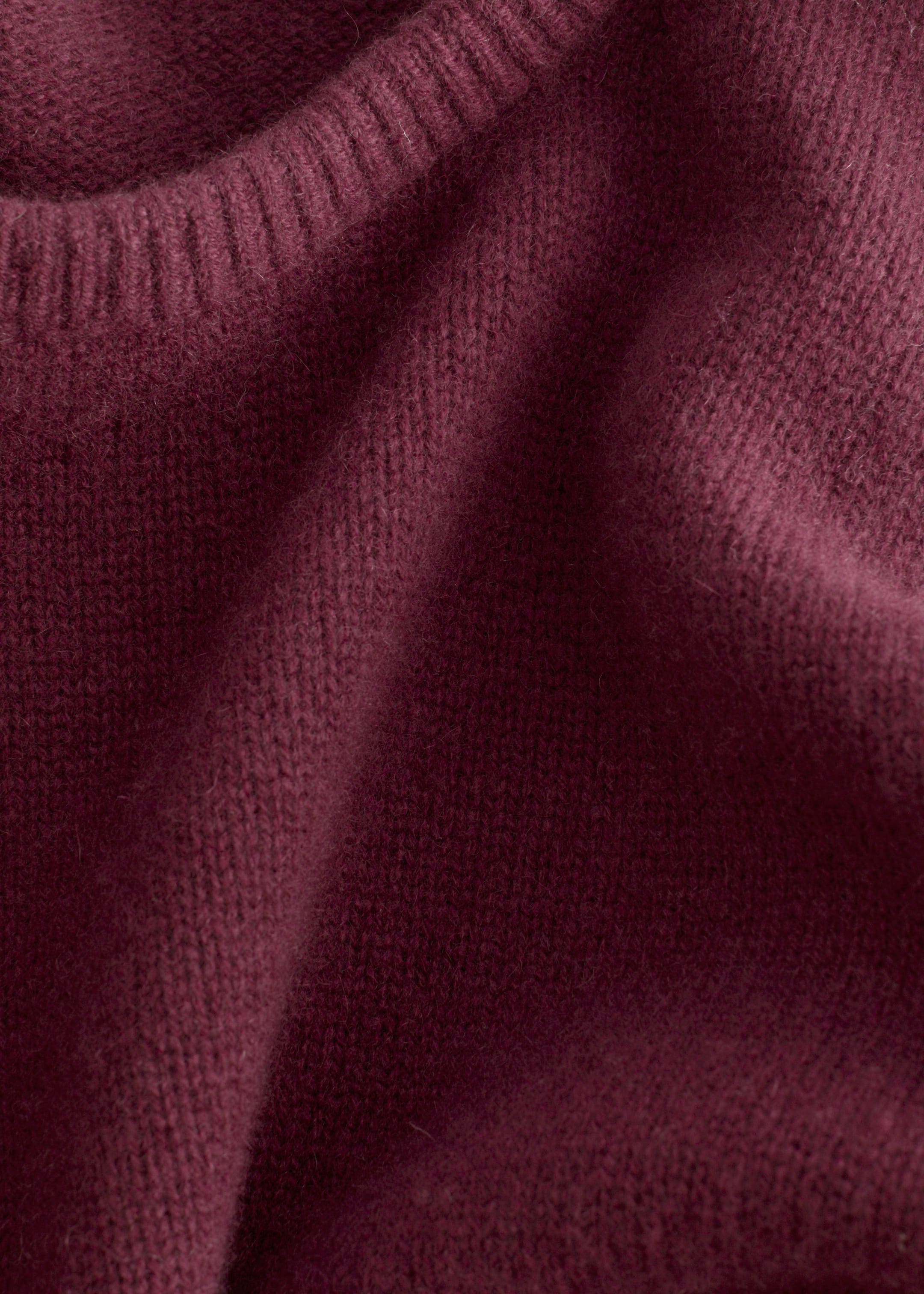 Grotere afbeelding bekijken: Een close-up toont een bordeauxrood gebreid T-shirt, waarbij de zachte, getextureerde kasjmier stof en de geribde afwerking langs de ronde hals worden benadrukt.