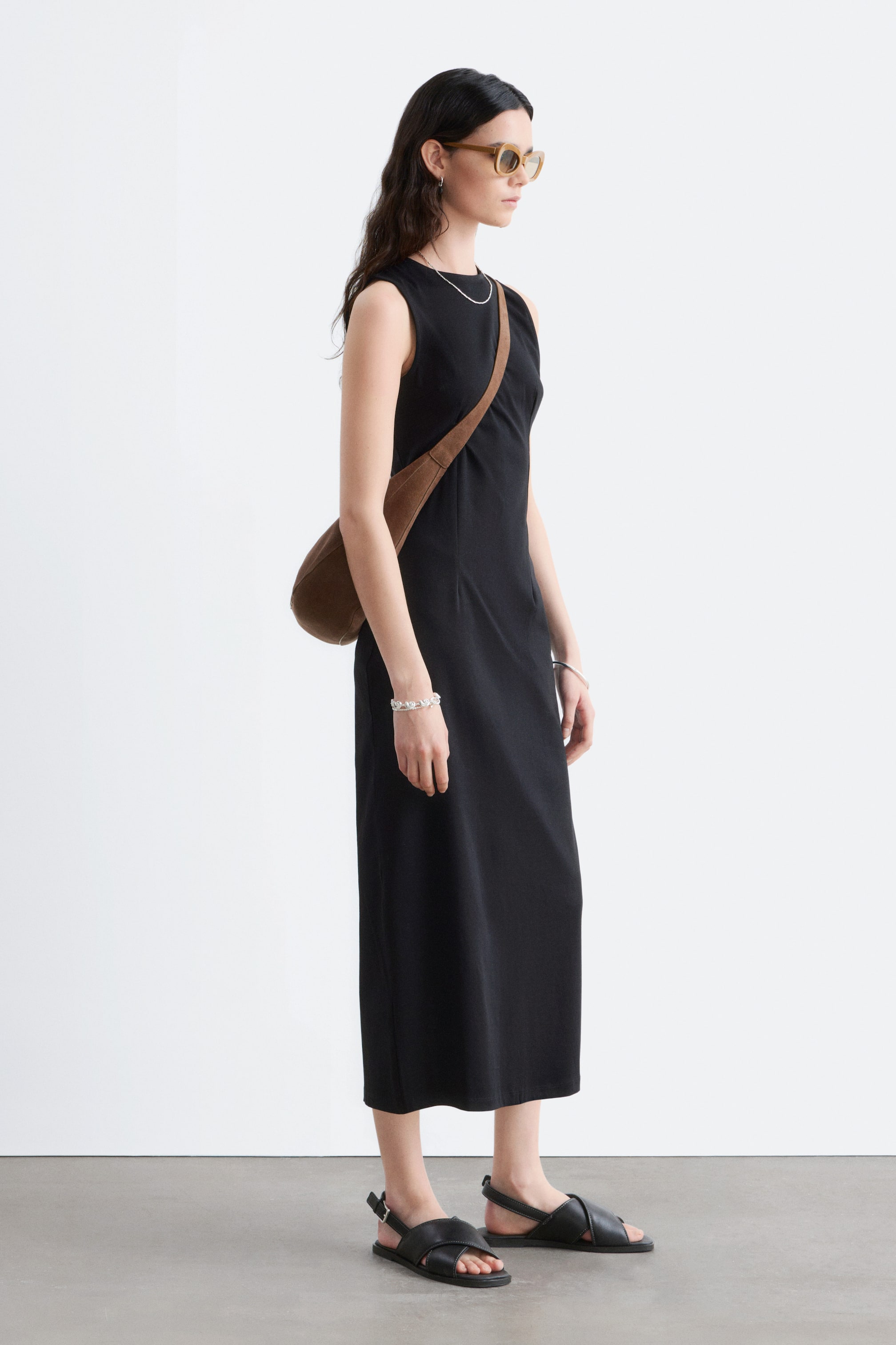 Agrandir l'image: Une robe midi noire sans manches présente un col rond et une silhouette droite, vue sous un léger angle latéral, avec une bandoulière marron traversant son devant.