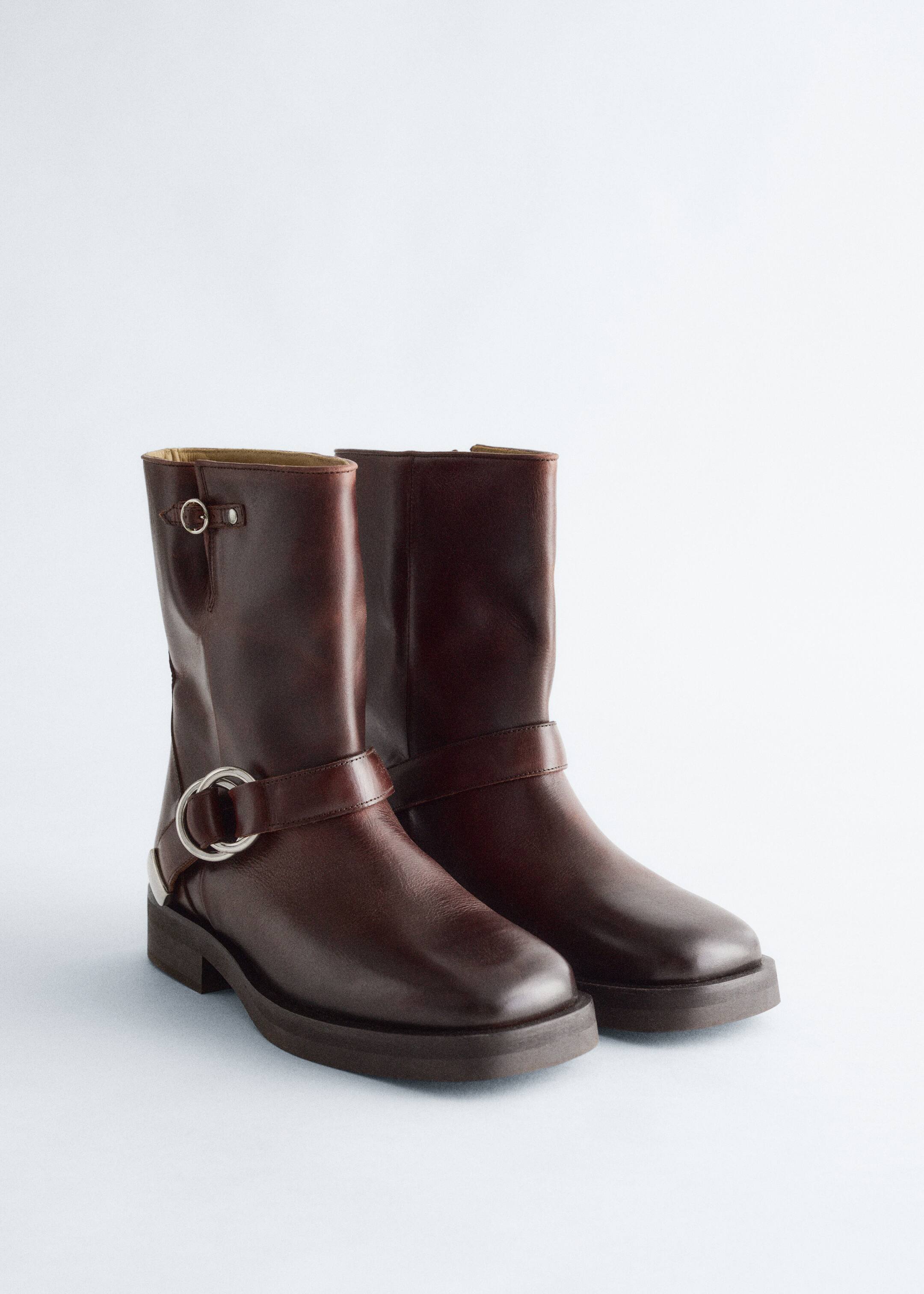 Größeres Bild anzeigen: Ein Paar dunkelbraune Lederstiefel im Biker-Stil steht in einem leichten Frontwinkel und zeigt klobige Sohlen und silberfarbene Metallbeschläge. Die Stiefel verfügen über eine kleine Schnalle nahe der Spitze des Schafts und eine große O-Ring-Schnalle mit einem Riemen, der den Knöchel umschließt.
