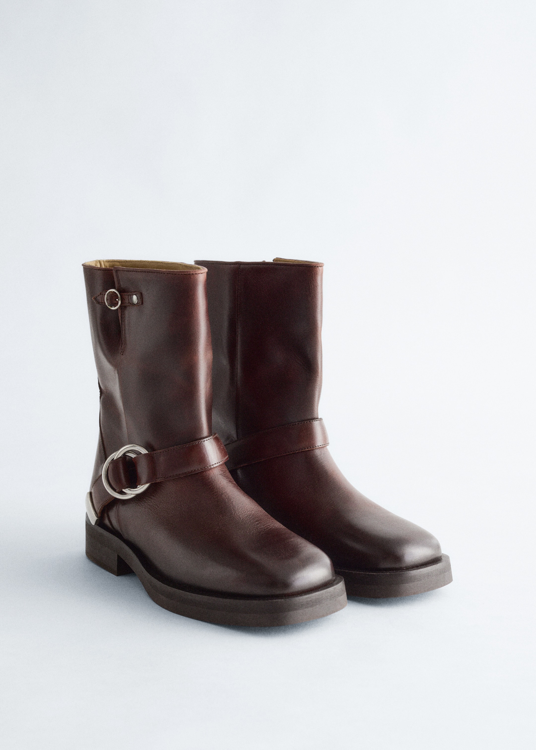 Größeres Bild ansehen: Ein Paar dunkelbraune Lederstiefel im Biker-Stil steht in einem leichten Frontwinkel und zeigt klobige Sohlen und silberfarbene Metallbeschläge. Die Stiefel verfügen über eine kleine Schnalle nahe der Spitze des Schafts und eine große O-Ring-Schnalle mit einem Riemen, der den Knöchel umschließt.