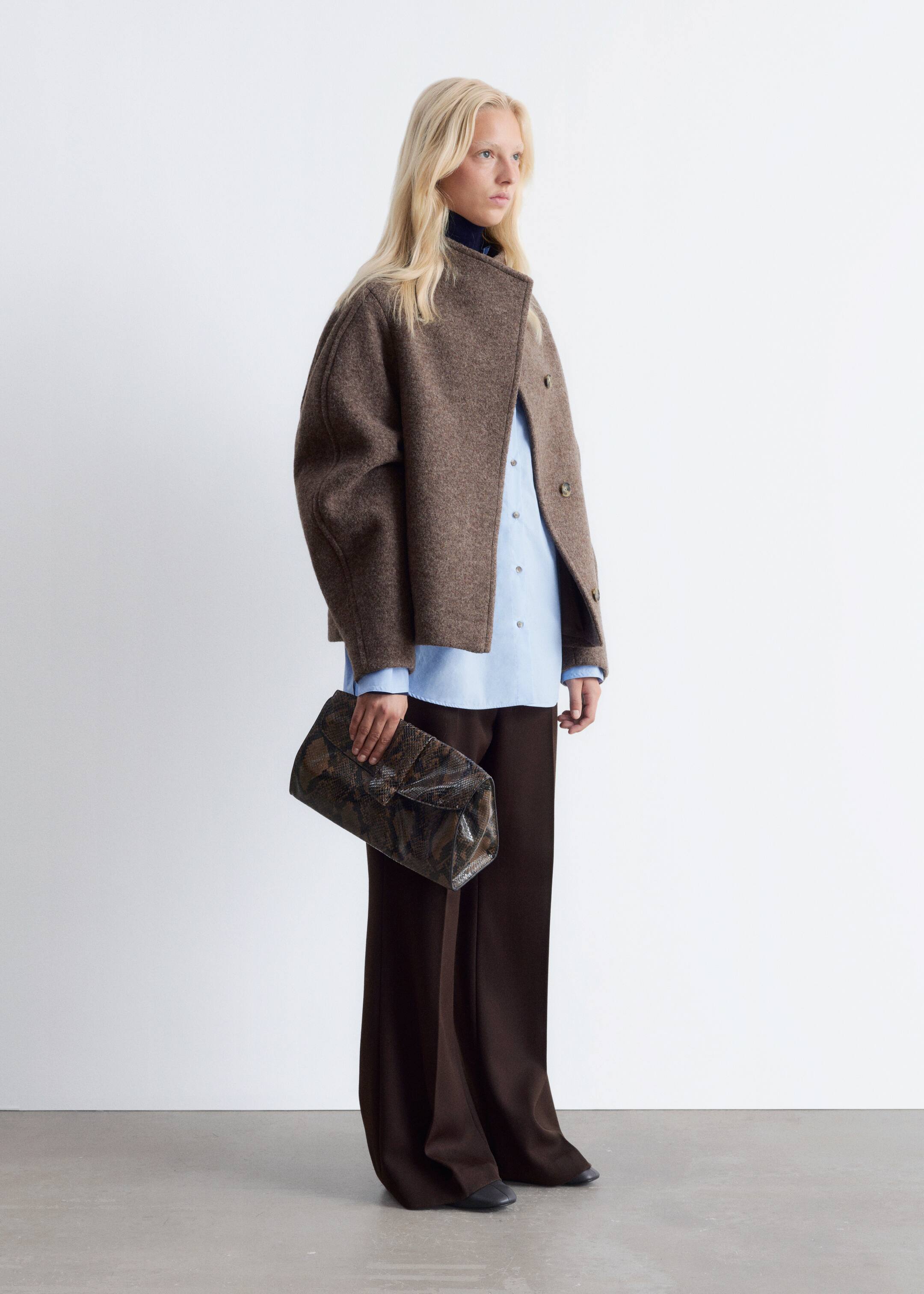 Grotere afbeelding bekijken: Een bruine, getextureerde korte jas met een opstaande kraag en knopen aan de voorkant wordt gedragen over een lichtblauw overhemd met knopen en donkerbruine wijde broek. Een rechthoekige clutch met een donker patroon aan de buitenkant wordt in één hand vastgehouden, wat de outfit compleet maakt.