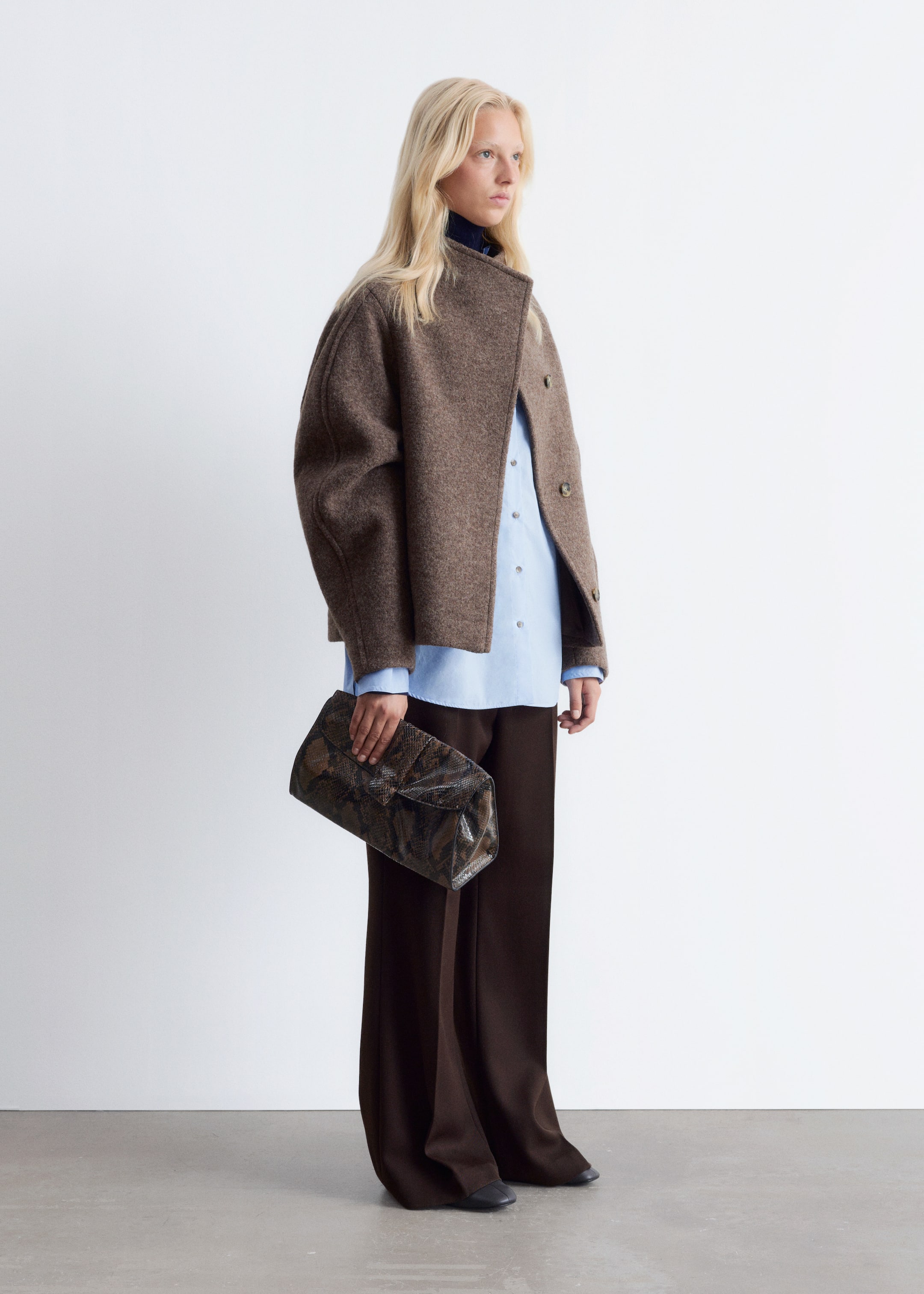 View larger image: A brown, textured short jacket featuring a stand collar and front buttons is being worn over a light blue button-down shirt and dark brown wide-leg pants. A rectangular clutch bag with a dark patterned exterior is being held in one hand, completing the outfit.