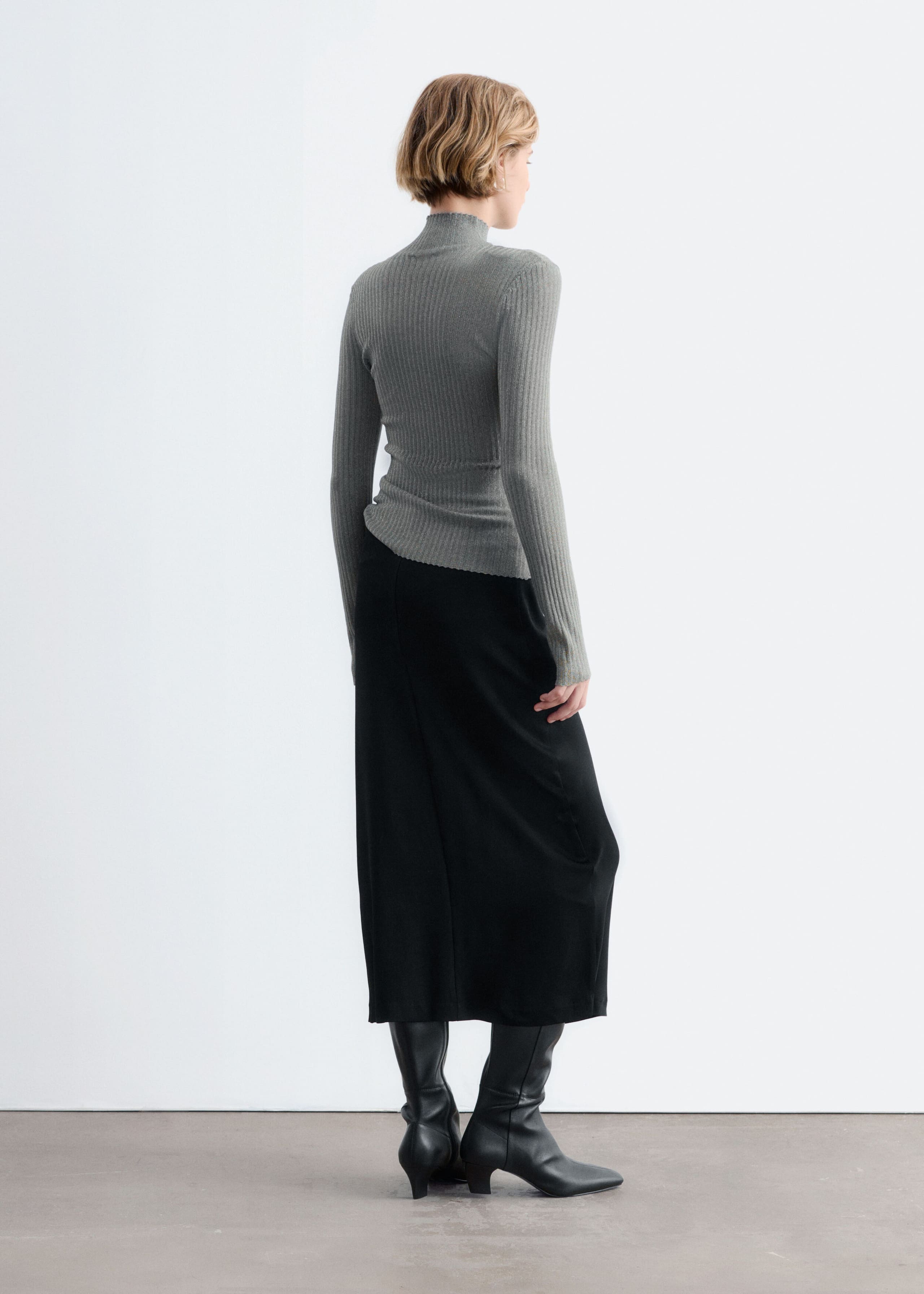 A grey, rib-knit mock neck top with a subtle metallic sheen is worn by the model, who is posed with their back partially turned. The long-sleeve top is styled with a black midi skirt and black leather heeled boots.
