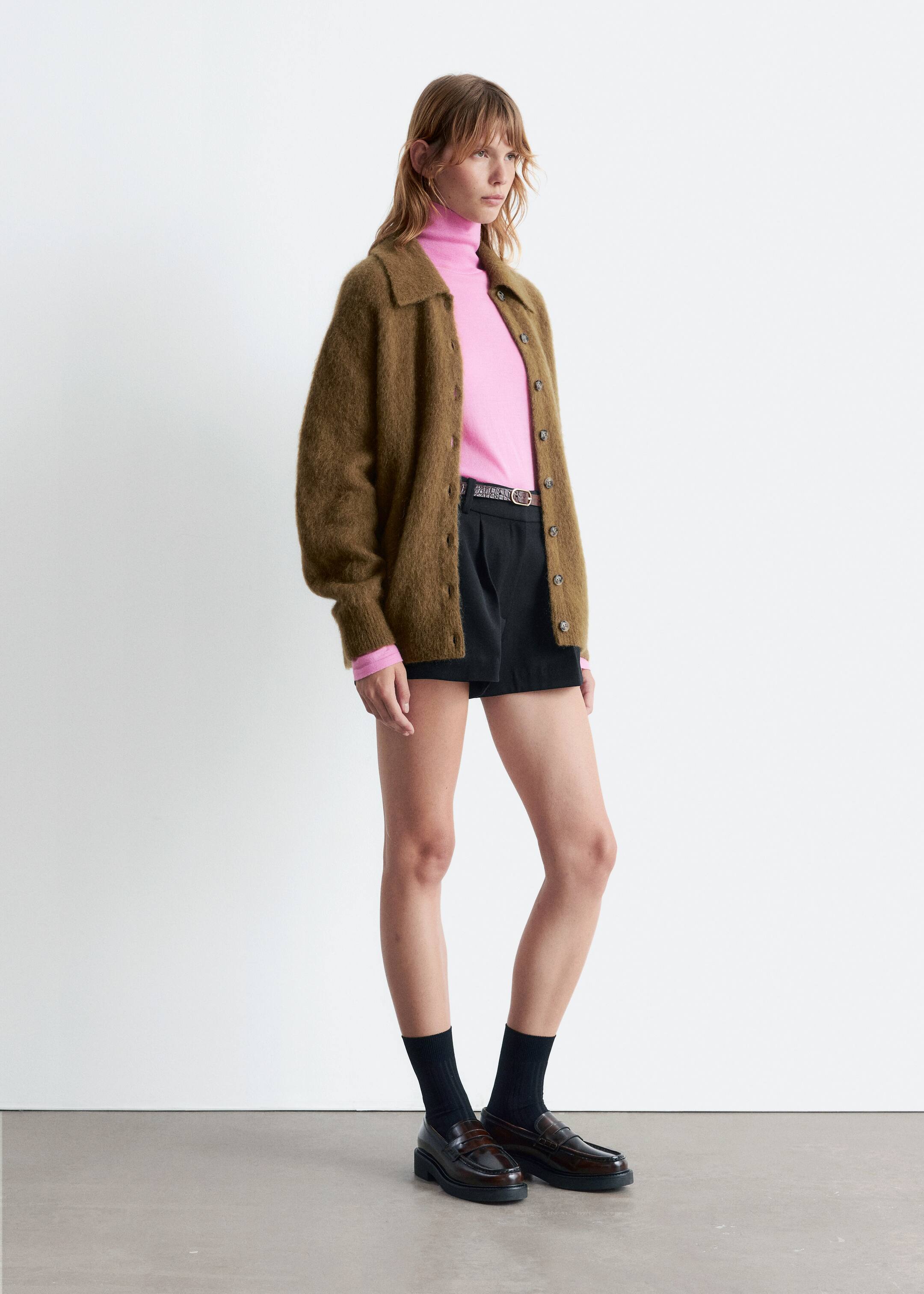 Größeres Bild ansehen: Ein Outfit wird im Profil gezeigt, nach rechts gerichtet. Es besteht aus einer olivbraunen, flauschigen Strickjacke mit Knopfleiste und Hemdkragen, die über einem rosa Rollkragenpullover getragen wird. Schwarze plissierte Shorts werden mit einem dünnen dunklen Gürtel, schwarzen gerippten Crew-Socken und dunkelbraunen klobigen Loafern getragen.