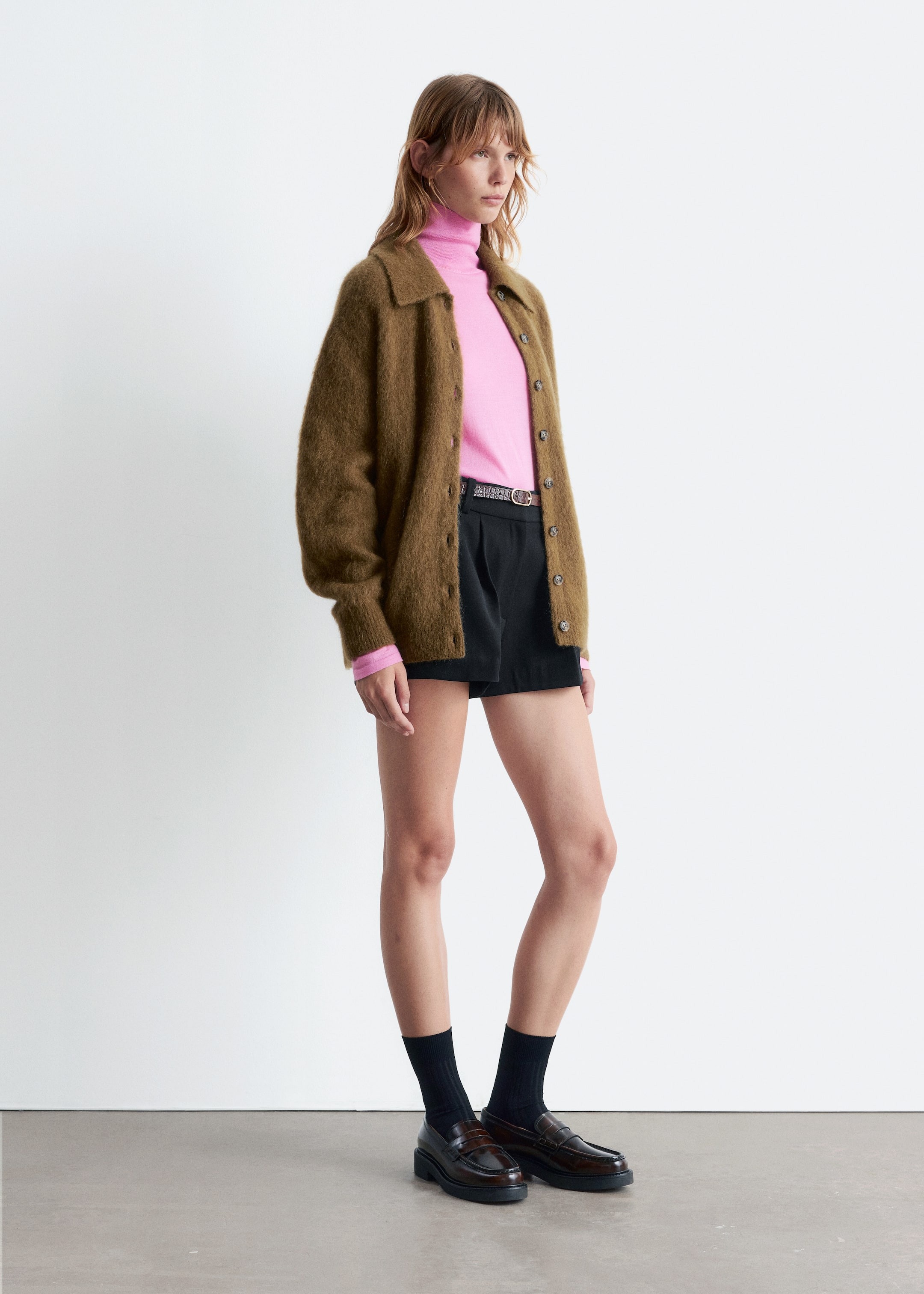 Grotere afbeelding bekijken: Een outfit wordt in profiel getoond, naar rechts gericht. Het bestaat uit een olijfbruine, pluizige cardigan met knoopsluiting en een overhemdkraag, die over een roze coltrui wordt gedragen. Zwarte geplooide shorts worden gedragen met een dunne donkere riem, zwarte geribbelde crew sokken en donkerbruine chunky loafers.
