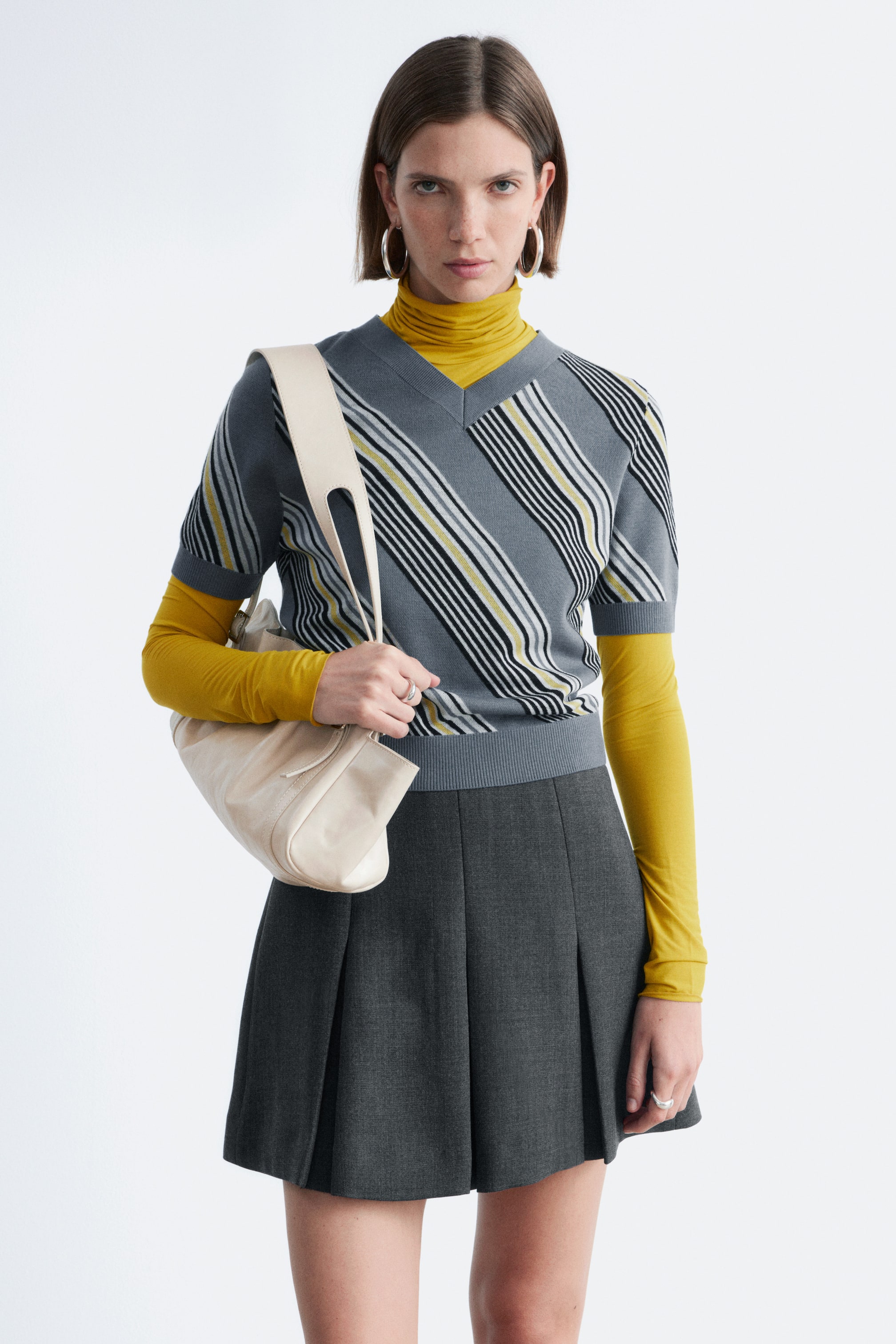Agrandir l'image: Un pull gris à col en V, avec des rayures diagonales noires, blanches et jaunes, des manches courtes et un ourlet côtelé, est porté par-dessus un col roulé jaune.