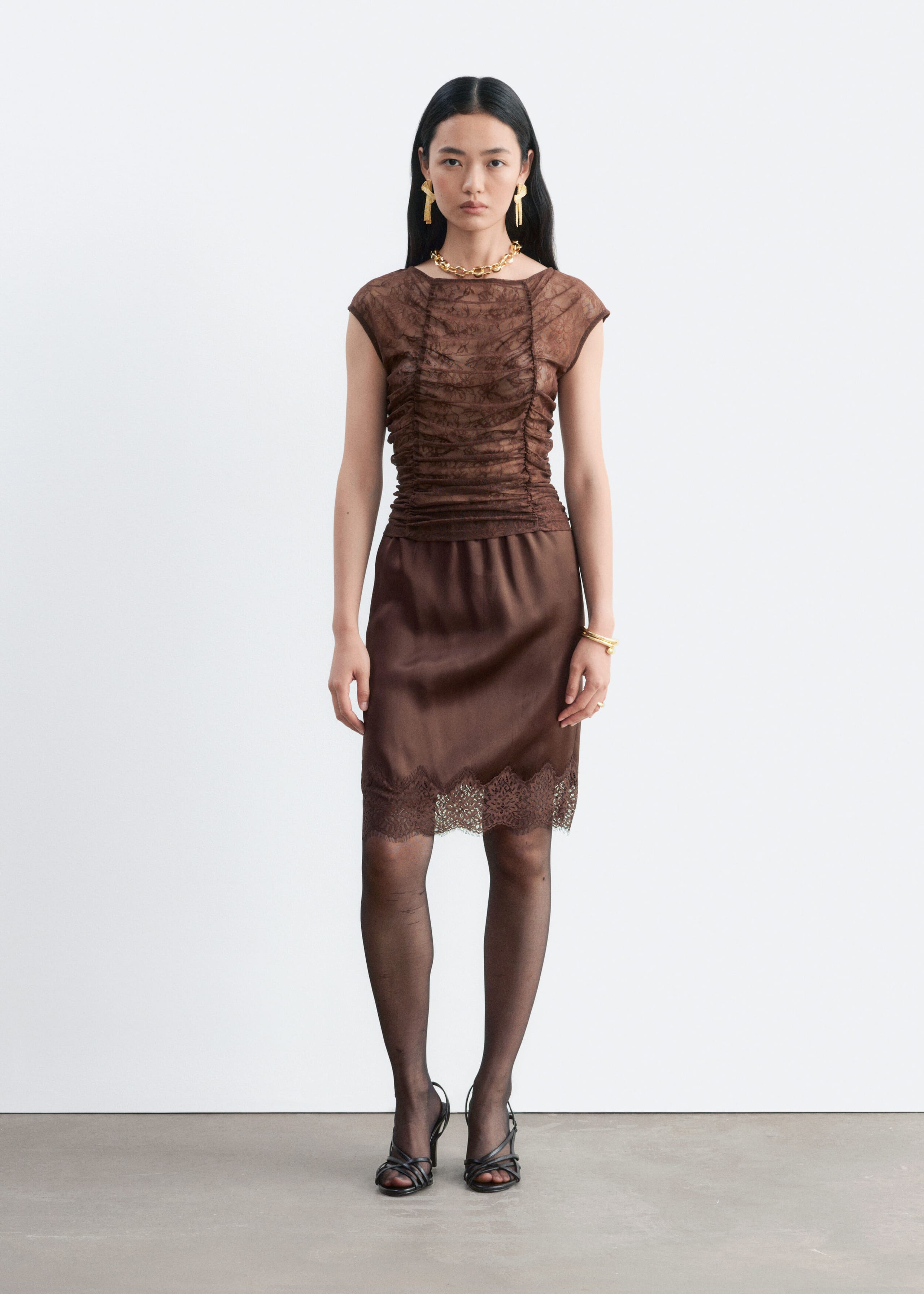 A brown lace top featuring ruched detailing through the body, cap sleeves, and a boat neckline is worn by the model. The top is paired with a matching brown satin skirt featuring a lace-trimmed hem.