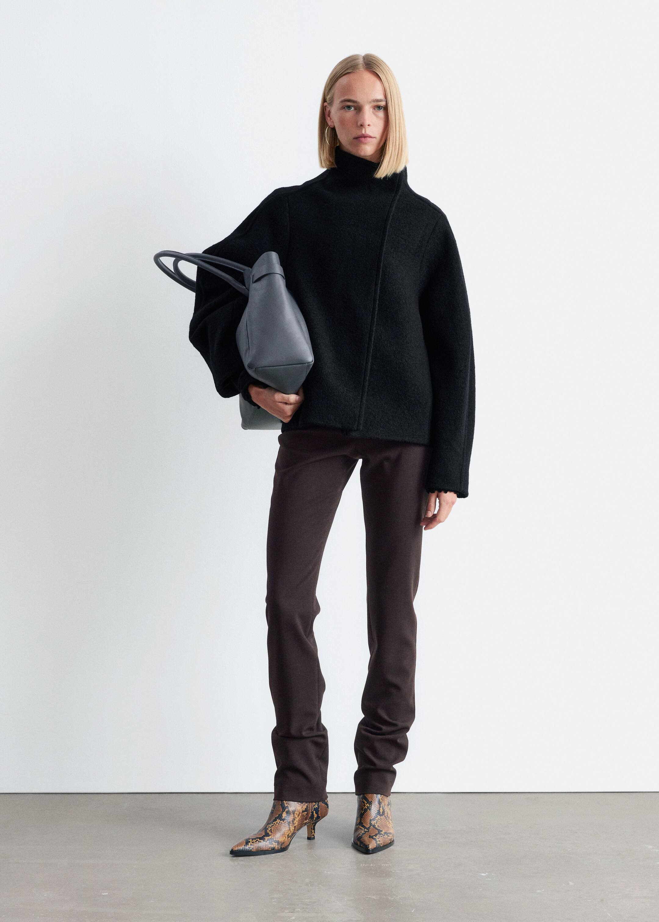 Visa större bild: En svart, texturerad stickad tröja med en hög trattkrage och en asymmetrisk framsöm bärs, med vida ärmar och åtsittande muddar. Mörkbruna, slim-fit byxor bärs också, som samlas vid anklarna, tillsammans med ormskinnsmönstrade stövlar med spetsiga tår och låga klackar. En mörkgrå strukturerad handväska med två topphandtag hålls, med alla produkter presenterade framåtvända och något vinklade.
