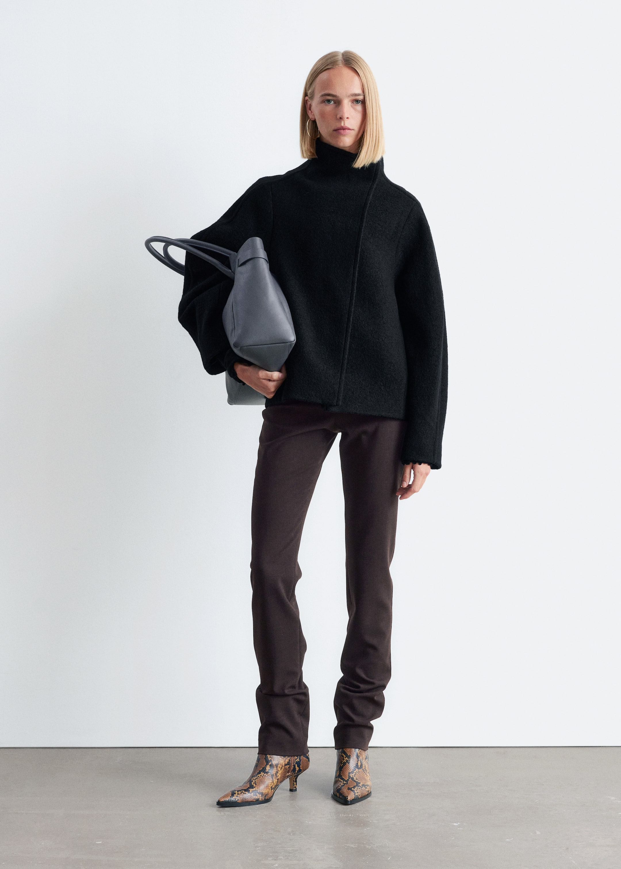 View larger image: A black, textured knit sweater with a high funnel neck and an asymmetrical front seam is being worn, featuring wide sleeves and fitted cuffs. Dark brown, slim-fit trousers are also being worn, pooling at the ankles, along with snakeskin-print boots with pointed toes and kitten heels. A dark gray structured tote bag with two top handles is being held, with all products presented front-facing and slightly angled.