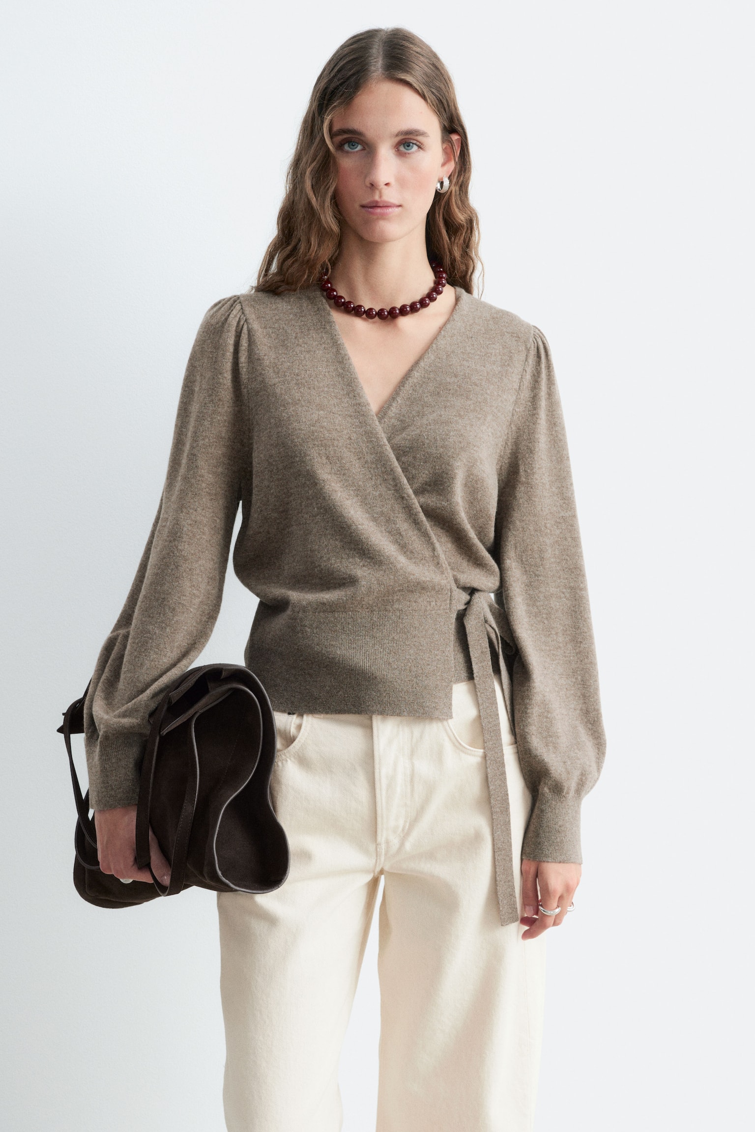 Una cardigan avvolgente in maglia color taupe-marrone è indossata dalla modella, con scollo a V, maniche lunghe a palloncino con polsini a coste e dettaglio da annodare lateralmente in vita. La modella abbina la cardigan a pantaloni color crema mentre tiene in mano una borsa in suede marrone scuro. - 1