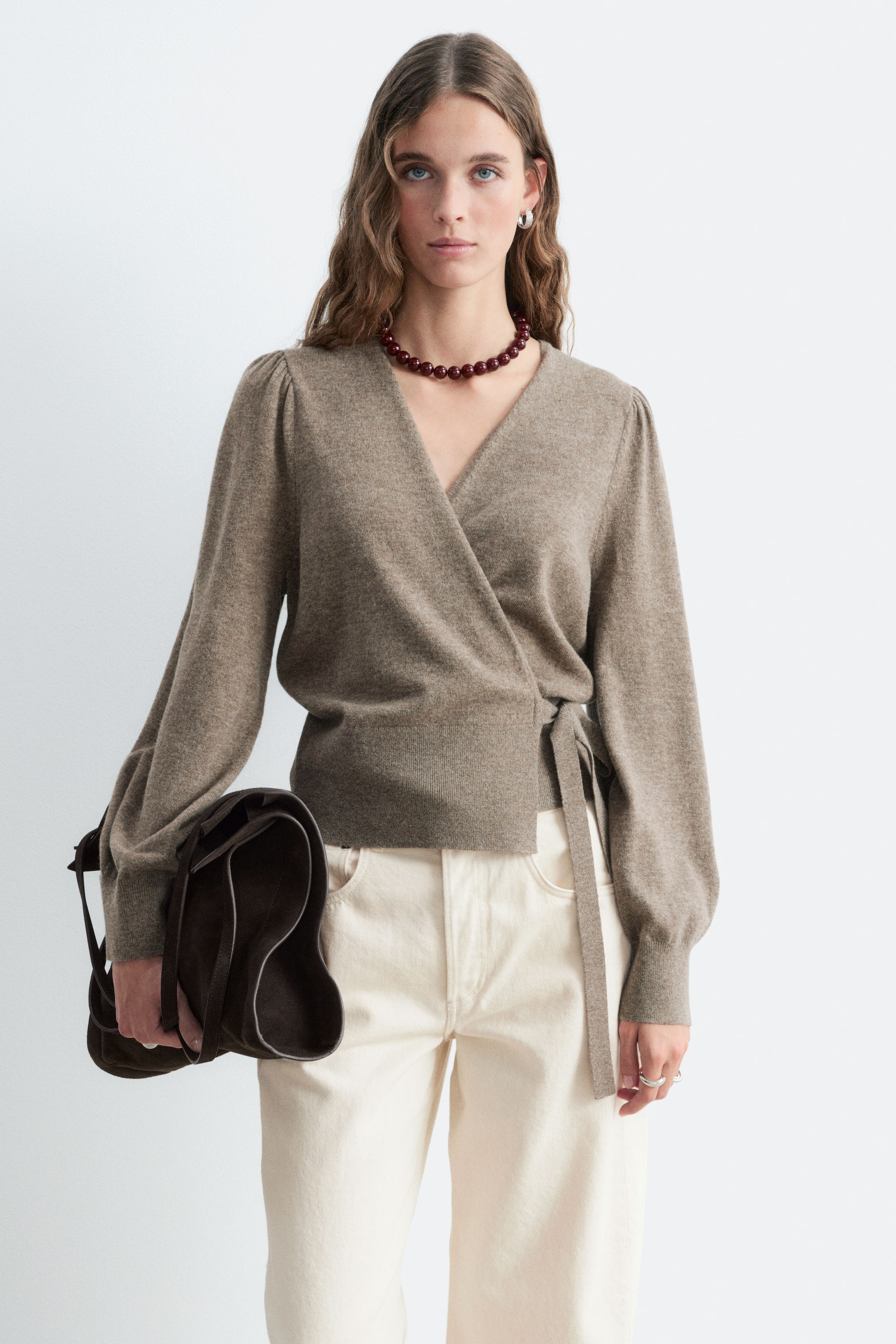 View larger image: The model is wearing a taupe-brown knitted wrap cardigan, featuring a V-neck, long puff sleeves with ribbed cuffs, and a self-tie detail at the side waist. The cardigan is styled with cream-coloured trousers and a dark brown suede bag.
