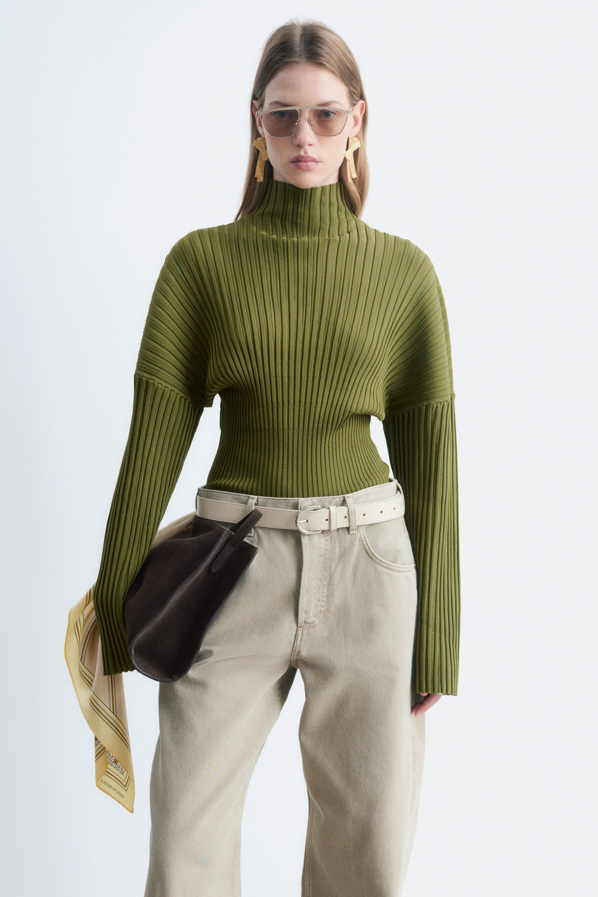 Agrandir l'image: Un pull vert à col montant en maille côtelée, dont les mailles se resserrent progressivement vers l’ourlet inférieur, est porté par le mannequin. Ce pull à coupe regular est associé à un pantalon clair et à une ceinture.