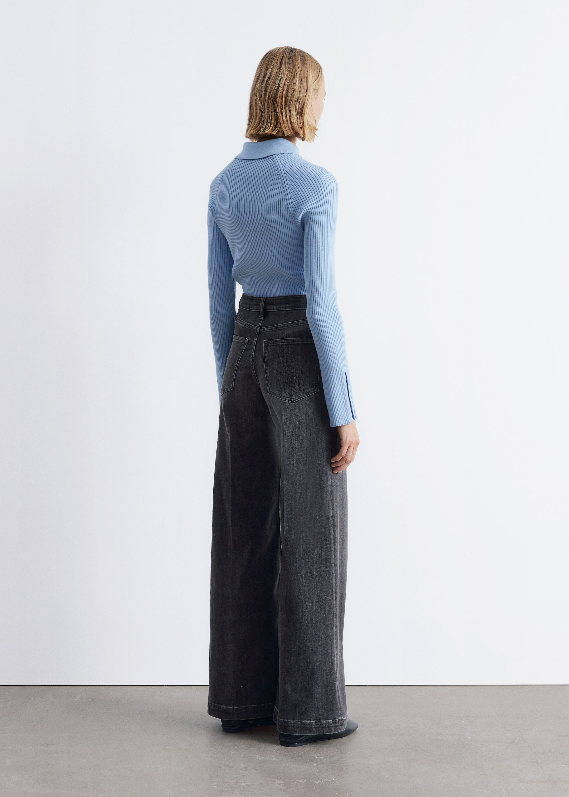 Visualizza immagine più grande: Un top a maglia a coste azzurro chiaro è indossato, caratterizzato da un colletto polo e maniche lunghe con sottili spacchi ai polsini. È abbinato a jeans grigio scuro a vita alta e gamba larga, che mostrano tasche posteriori applicate e una vestibilità ampia e rilassata sul retro.