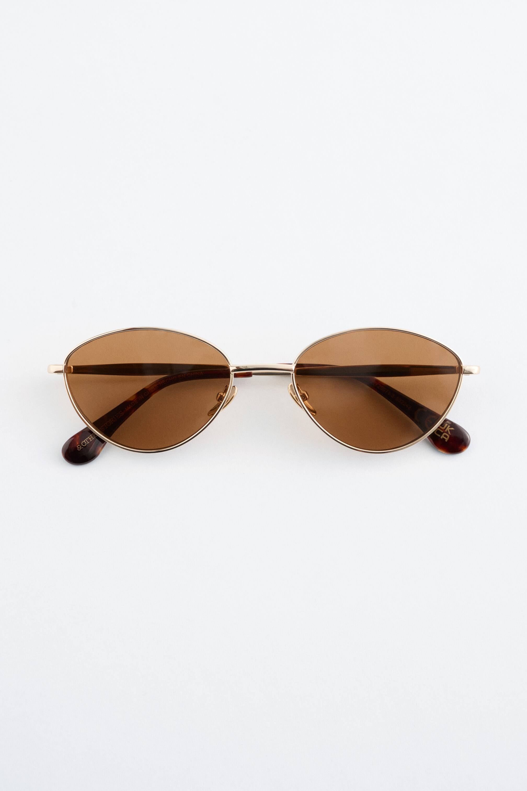 Преглед на по-голямо изображение: Слънчеви очила със златна рамка са поставени плоско, представяйки фронтален изглед. Те са с овални, кафяво оцветени лещи и тънки златни дръжки, които се простират до накрайници с костенурков ефект.