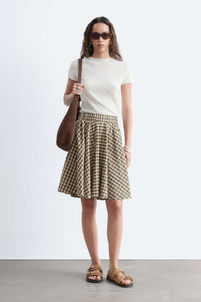 La falda presenta un patrón de cuadros en verde oliva, beige y blanco, con una cintura elástica, mostrando una silueta en línea A que cae por encima de la rodilla. - 1
