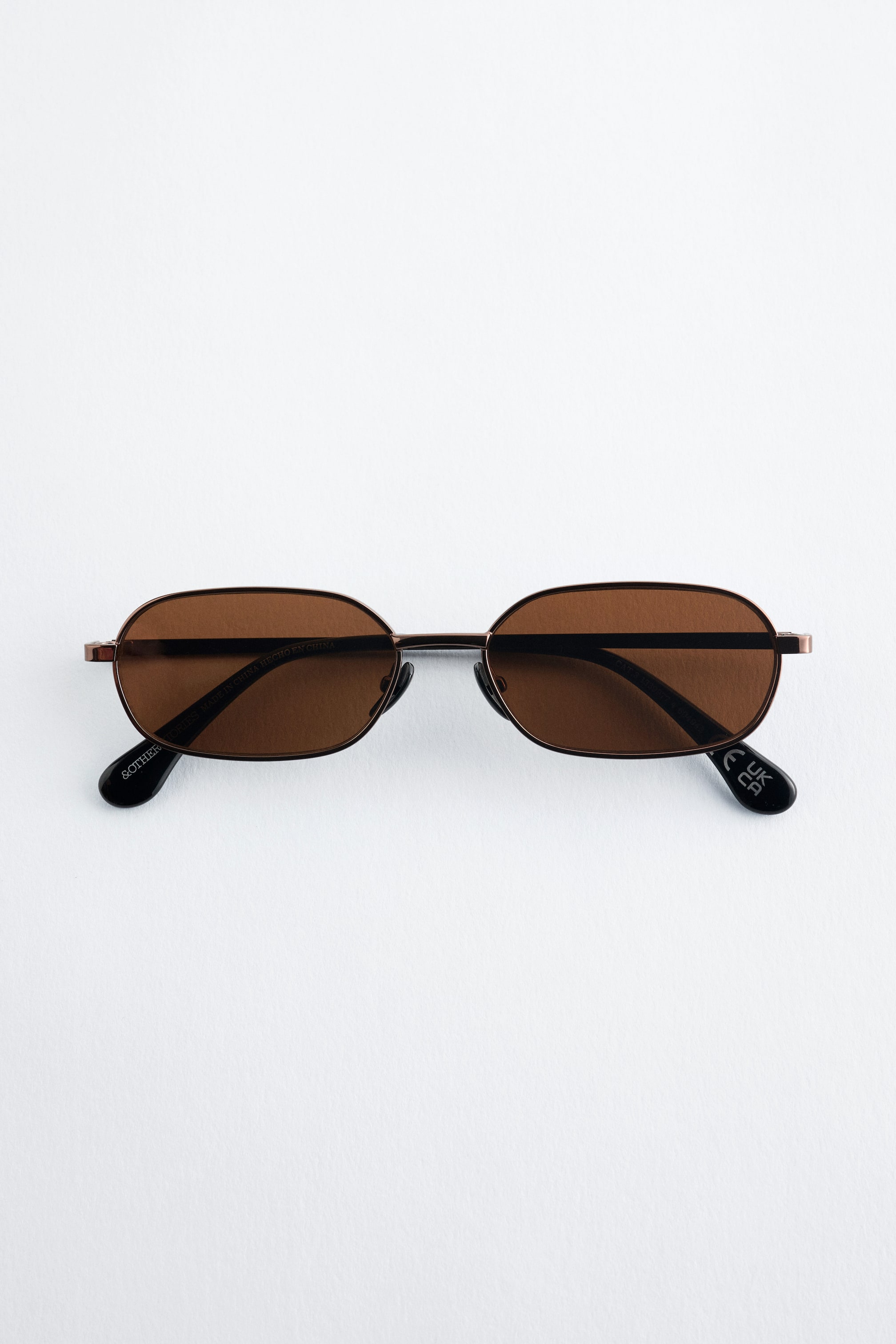 Ver imagen más grande: Las gafas de sol presentan una montura de acero inoxidable de tono bronce y diseño delgado. Muestran lentes pequeñas y rectangulares con un tinte marrón y bordes suavemente redondeados. Las gafas de sol se presentan desde una vista frontal.