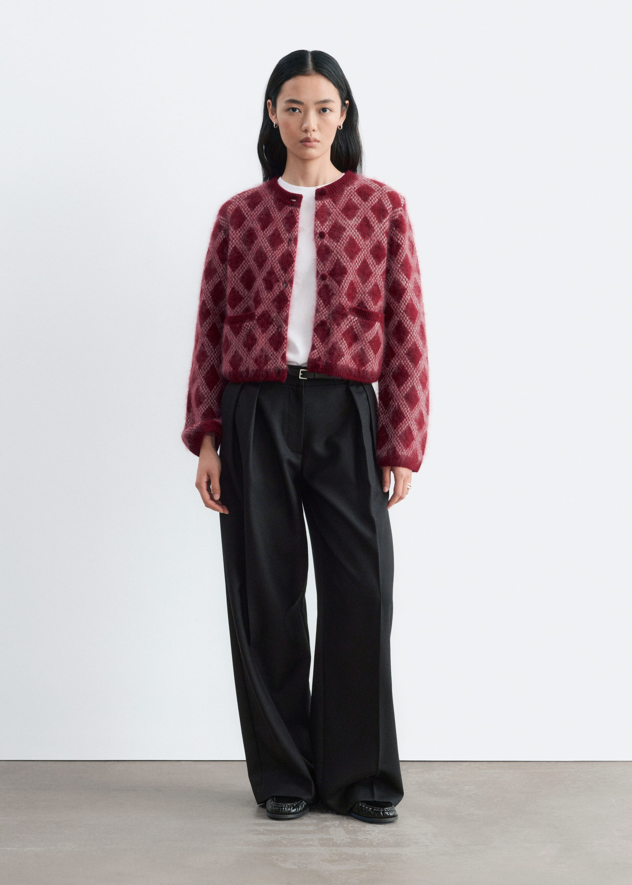 A fuzzy red cardigan featuring a light pink diamond pattern and structured shoulders is worn by the model over a white T-shirt and black wide-leg trousers. The cardigan is designed with a round neck, long sleeves, and a button-front closure.