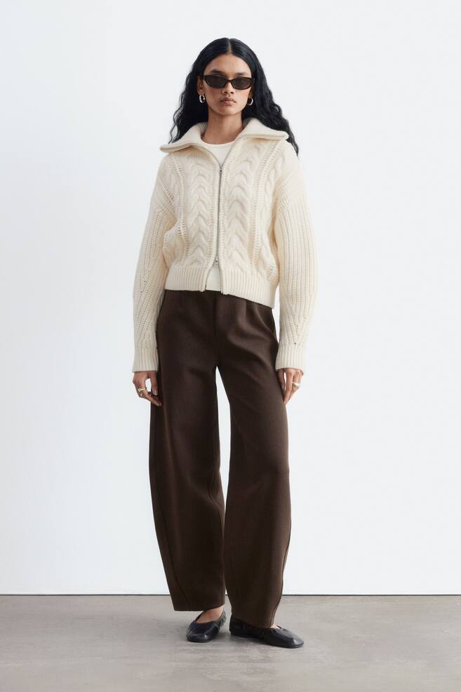 Dark brown scuba jersey trousers are worn by the model, featuring a high-rise waist with darts and wide, structured legs with curved side seams. The trousers are paired with a cream-coloured cable-knit zip-up sweater and black flat shoes. - 1