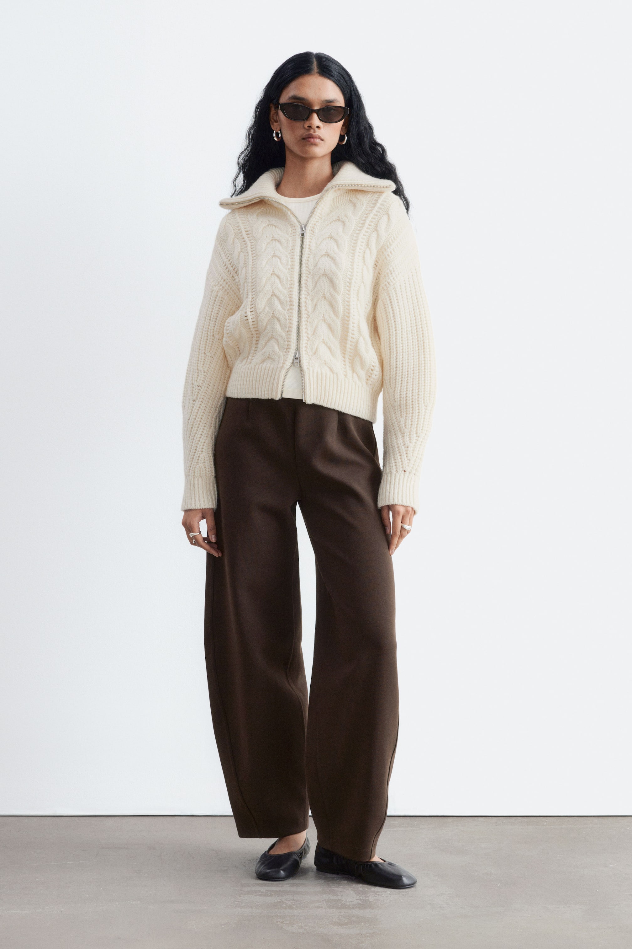 View larger image: Dark brown scuba jersey trousers are worn by the model, featuring a high-rise waist with darts and wide, structured legs with curved side seams. The trousers are paired with a cream-coloured cable-knit zip-up sweater and black flat shoes.