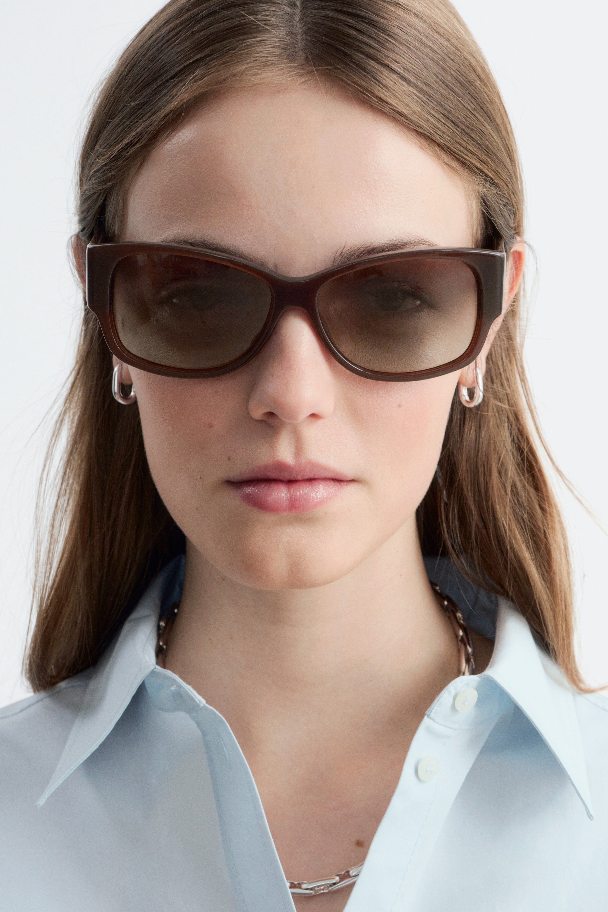 Agrandir l'image: Lunettes de soleil en acétate marron, avec une monture ovale aux bords arrondis, des branches larges et des verres teintés ton sur ton, portées par le mannequin. Les lunettes de soleil sont assorties à une chemise à col bleu clair et des bijoux en argent.