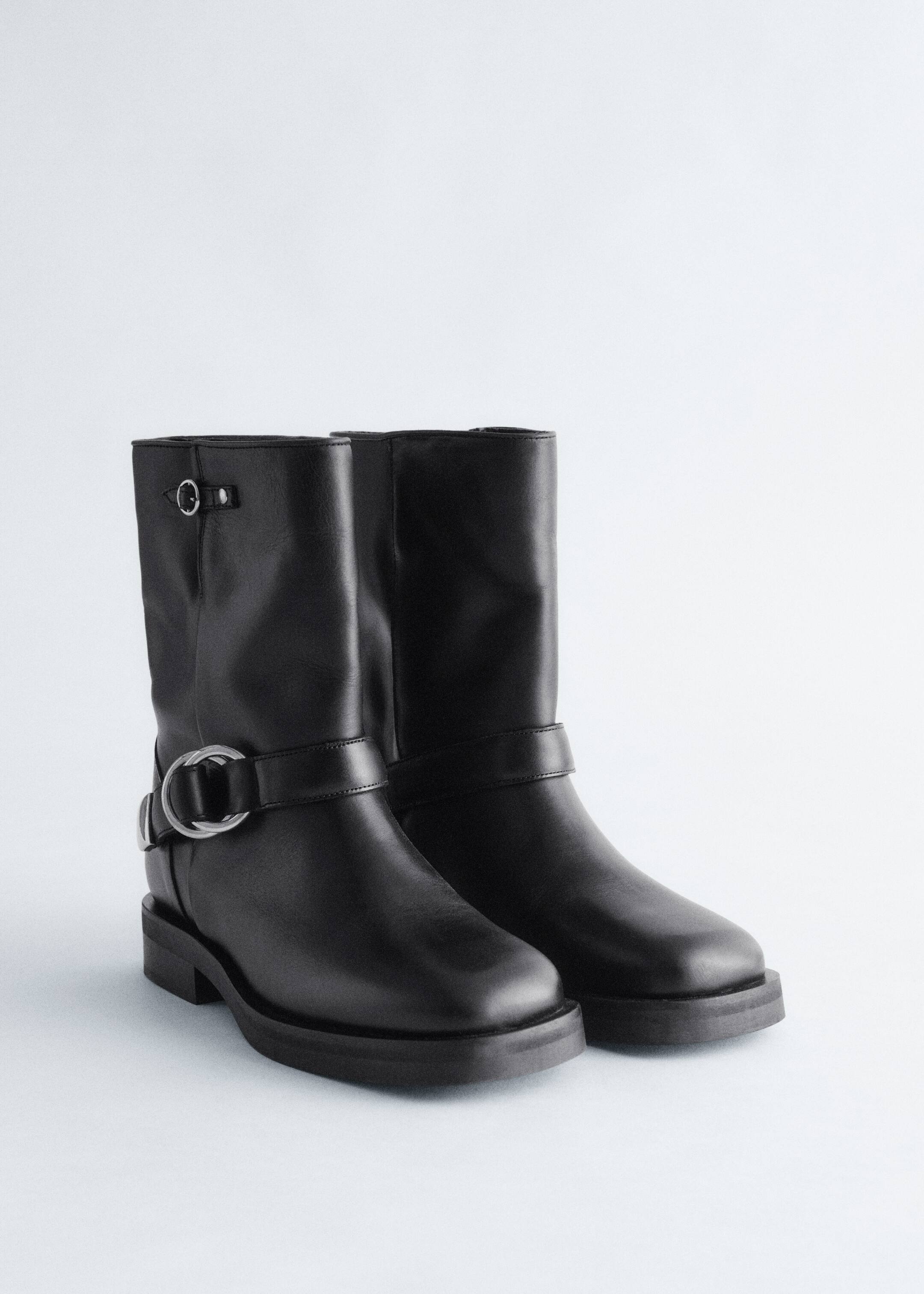Agrandir l'image: Une paire de bottes en cuir noir est posée à un léger angle, mettant en valeur des semelles épaisses et des éléments métalliques argentés d'inspiration biker, y compris une large sangle avec une grande boucle en D autour de la cheville de la botte gauche et un petit détail en anneau près du haut de sa tige.
