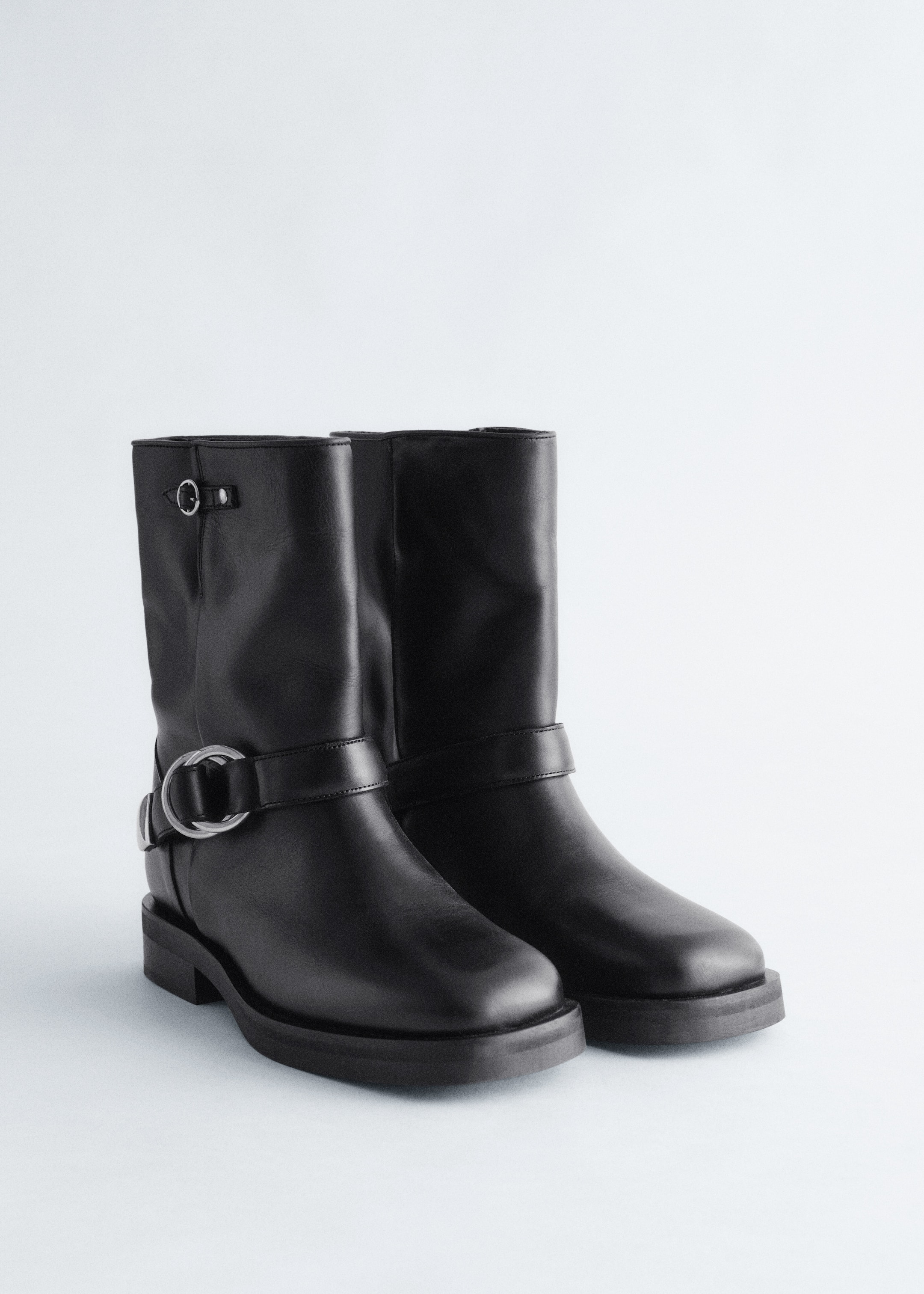 View larger image: A pair of black leather boots is standing at a slight angle, showcasing chunky soles and silver-toned biker-inspired hardware including a wide strap with a large D-ring buckle around the ankle of the left boot and a small ring detail near the top of its shaft.